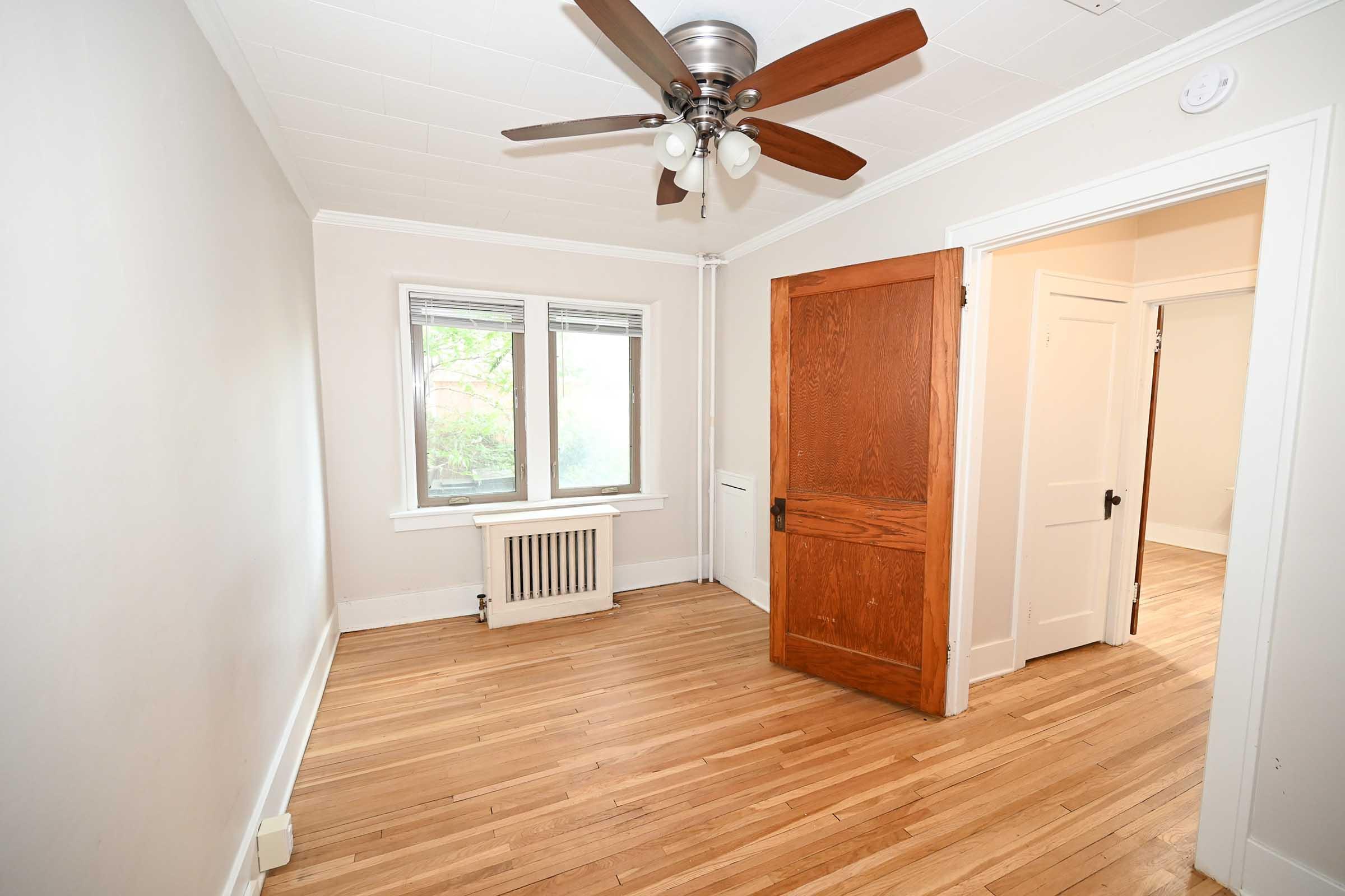 Empty room with hardwood flooring, featuring a ceiling fan, three windows allowing natural light, and an open wooden door leading to another space. The walls are painted in a light color, and there is a radiator near the window. The room has a simple and clean aesthetic.