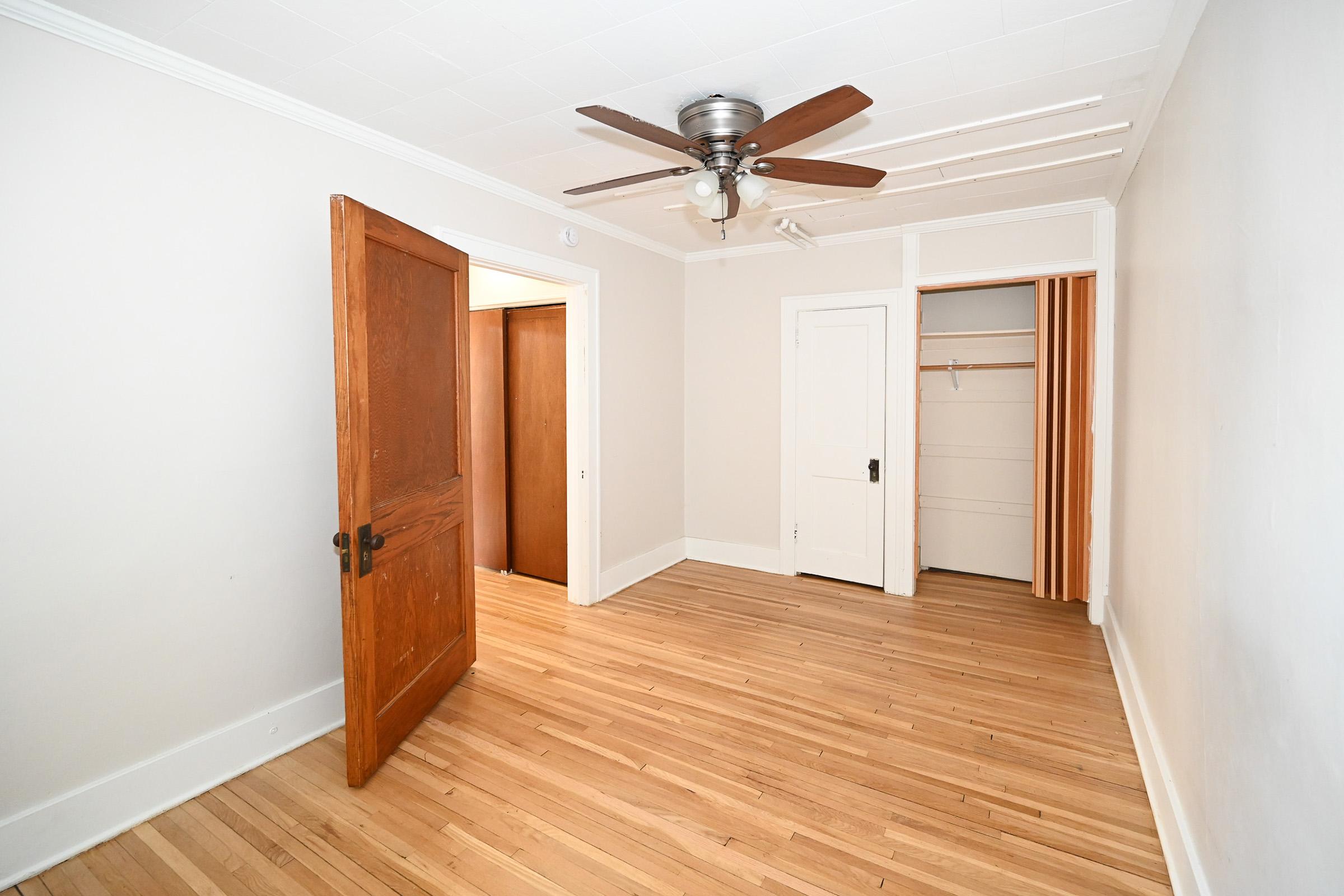 A spacious room with hardwood flooring, featuring an open door leading to a hallway and a closed door to a closet. The ceiling has a ceiling fan, and there's a smooth, light-colored wall. Natural light illuminates the area, creating a warm and inviting atmosphere.