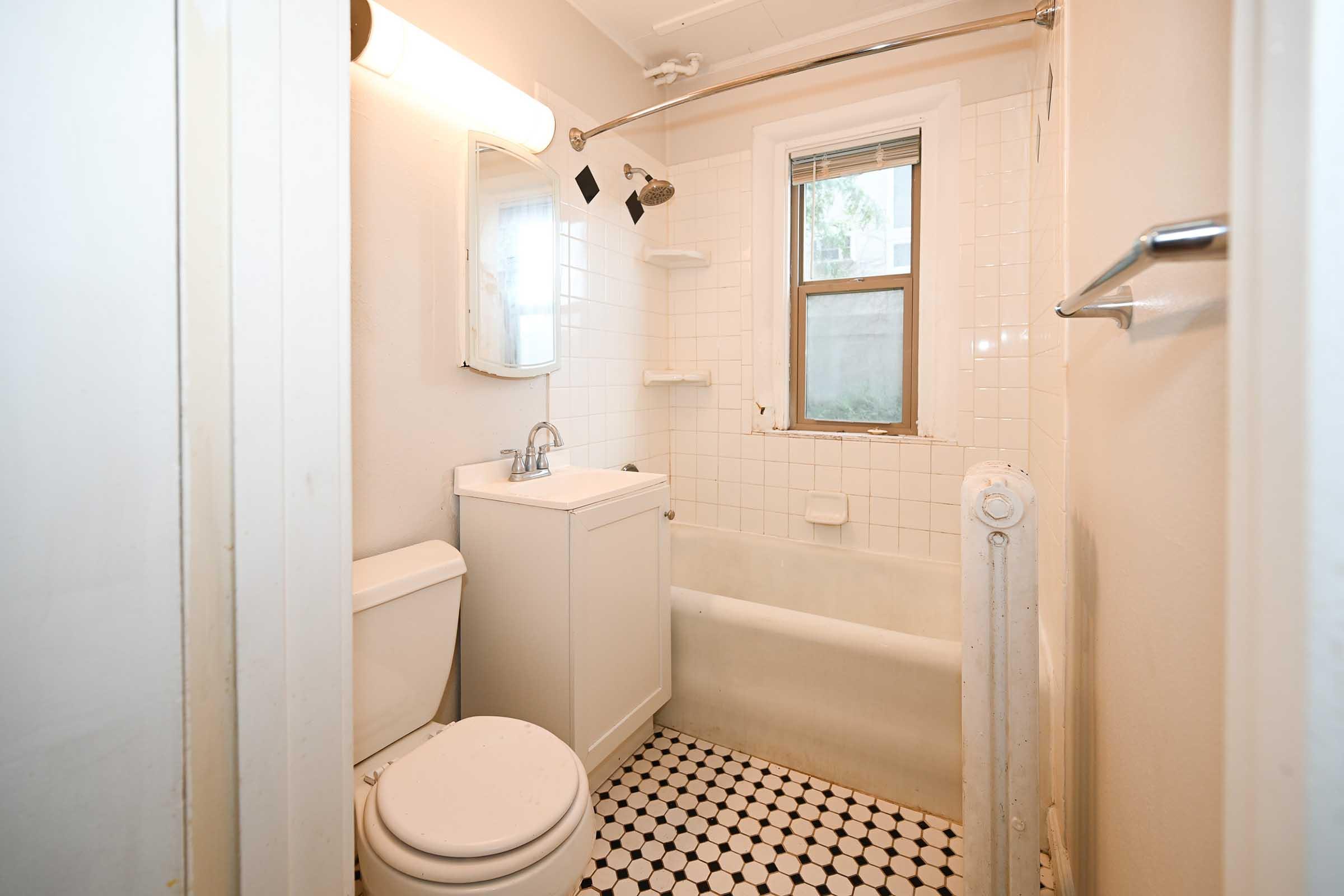A small bathroom featuring a white tiled shower with a glass door, a compact vanity with a sink, a toilet, and a mirror. The floor is covered with black and white patterned tiles. Natural light enters through a window, and there is a towel rack on the wall.