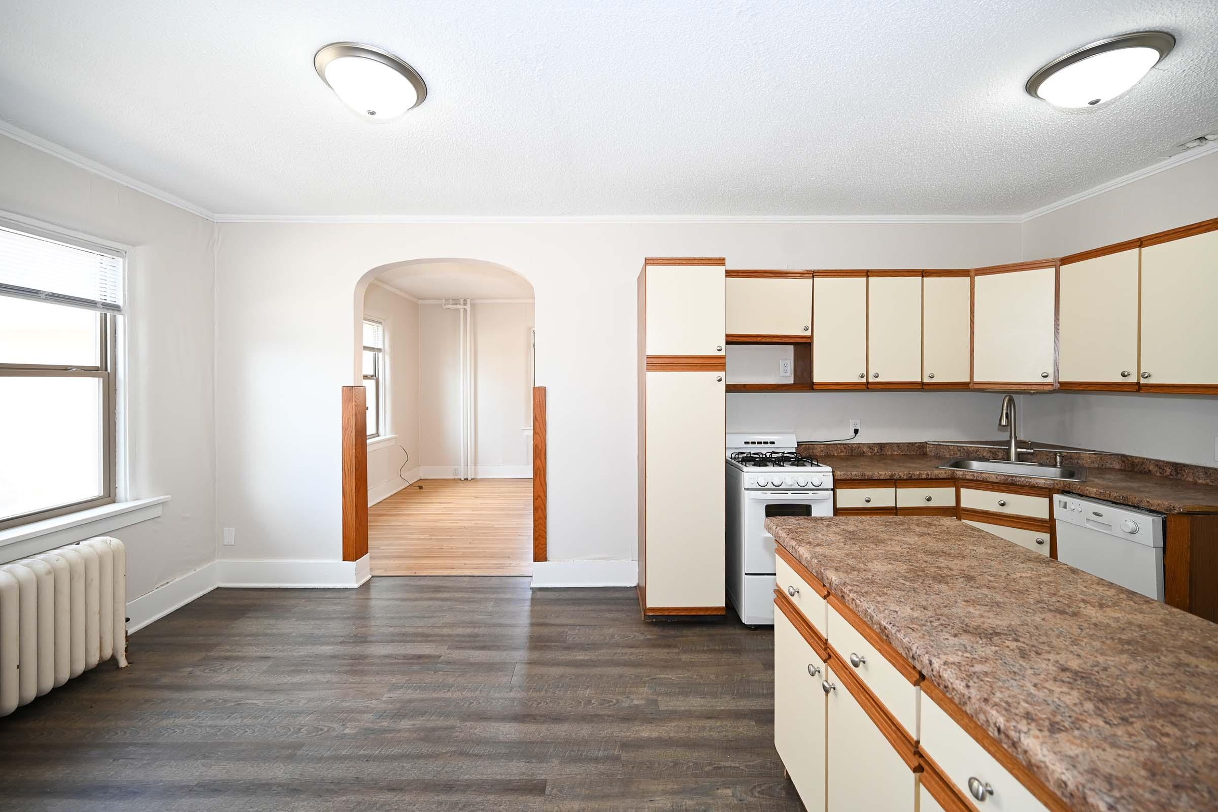 A bright, spacious kitchen featuring beige cabinets and a granite countertop. Includes a white stove and dishwasher, with an arched entry leading to another room. The hardwood floor adds warmth, and there's a vintage radiator on the side. Natural light streams in through the window.