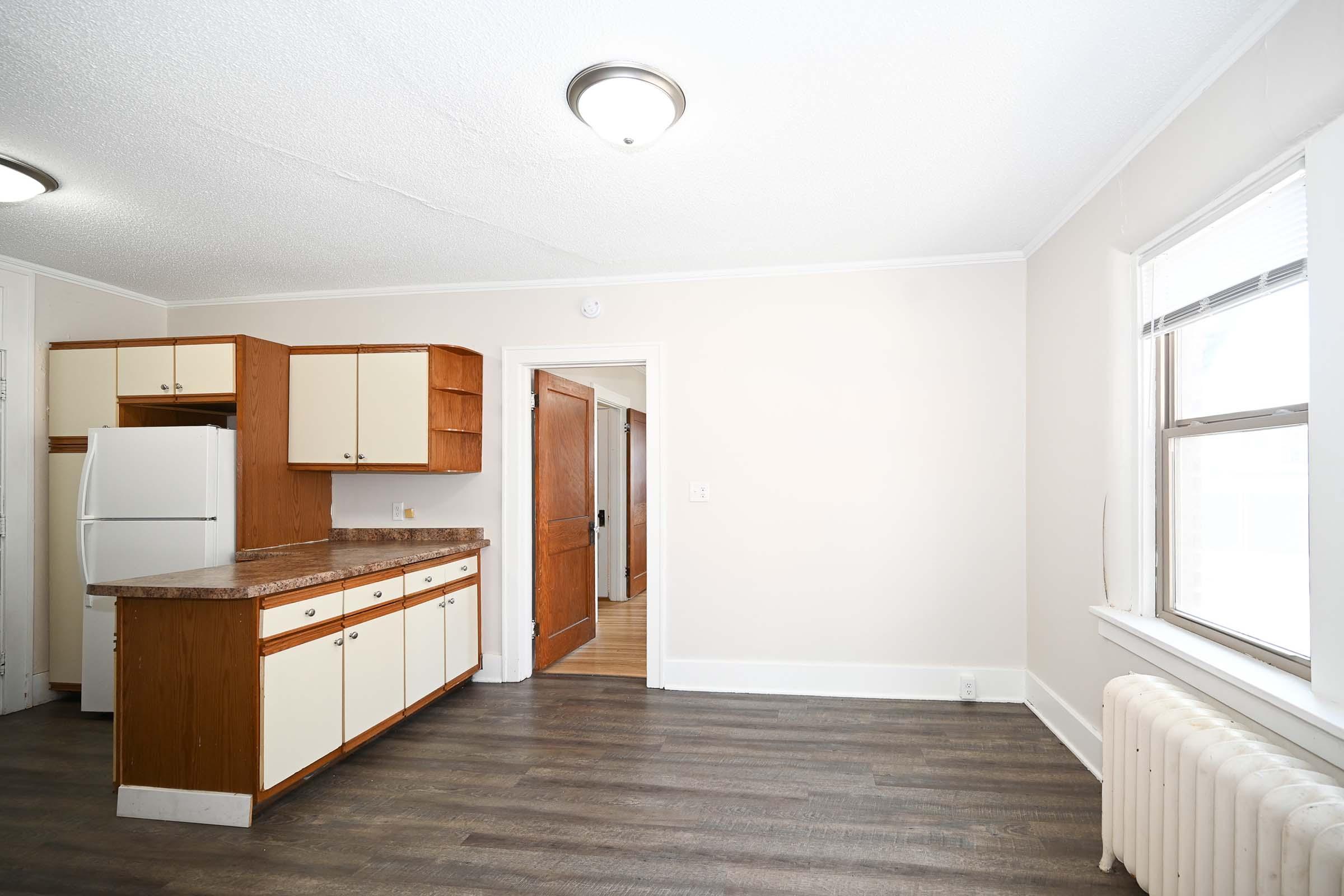 A bright kitchen featuring wooden cabinets, a marble countertop, and a white refrigerator. The space has a light-colored wall, a hardwood floor, and a window allowing natural light in. There's a door leading to another room, and a radiator is visible along the wall.