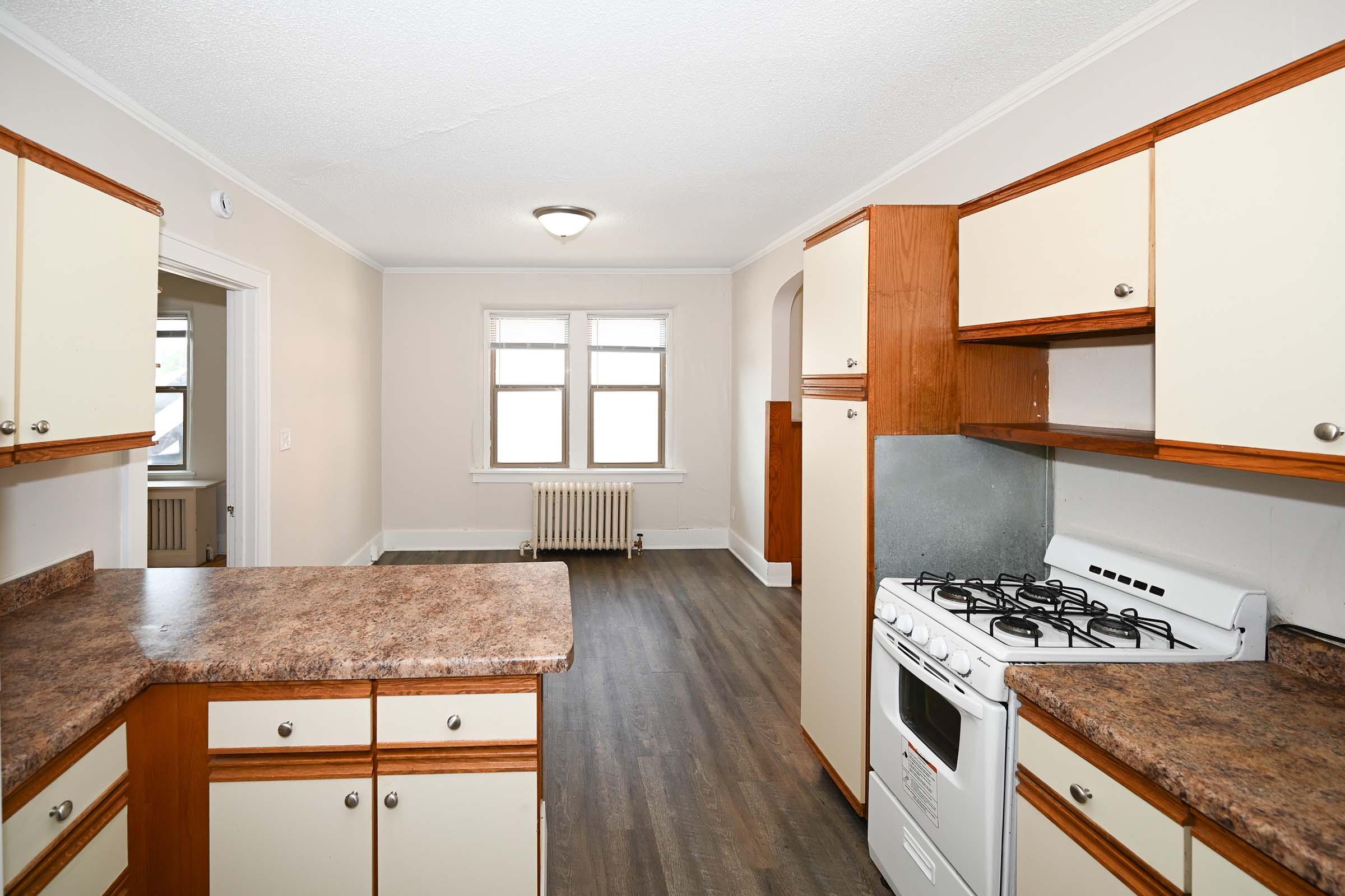 A spacious kitchen featuring wooden cabinets with cream-colored doors, a gas stove, and a countertop. The kitchen opens into a well-lit living area with large windows and hardwood flooring. An arched doorway leads to another room in the background.