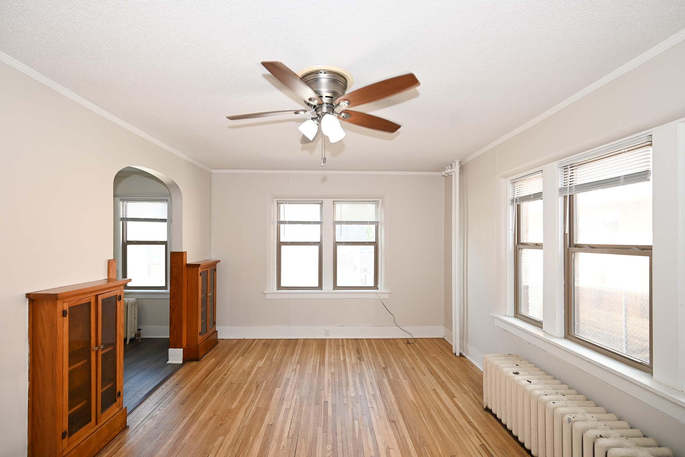A spacious, empty living room featuring hardwood floors, two large windows allowing natural light, and a ceiling fan. There are two wooden cabinets on either side of an arched entryway, and a radiator along the wall, creating a cozy atmosphere.
