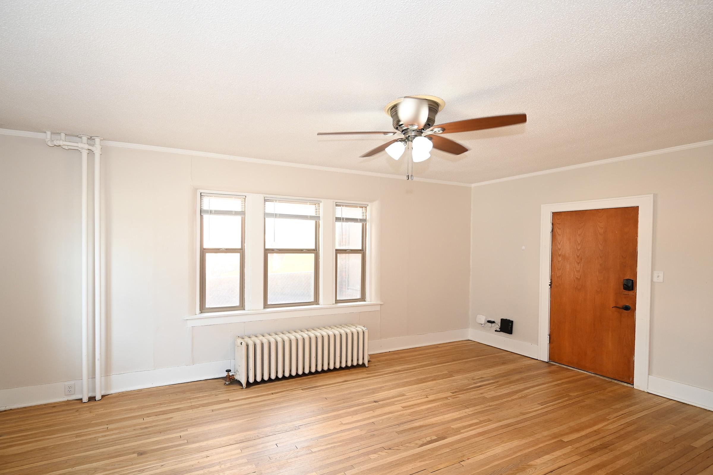 Spacious living room with hardwood floors, a ceiling fan, large windows letting in natural light, a radiator, and a wooden front door. The walls are painted a light color, creating an airy atmosphere.