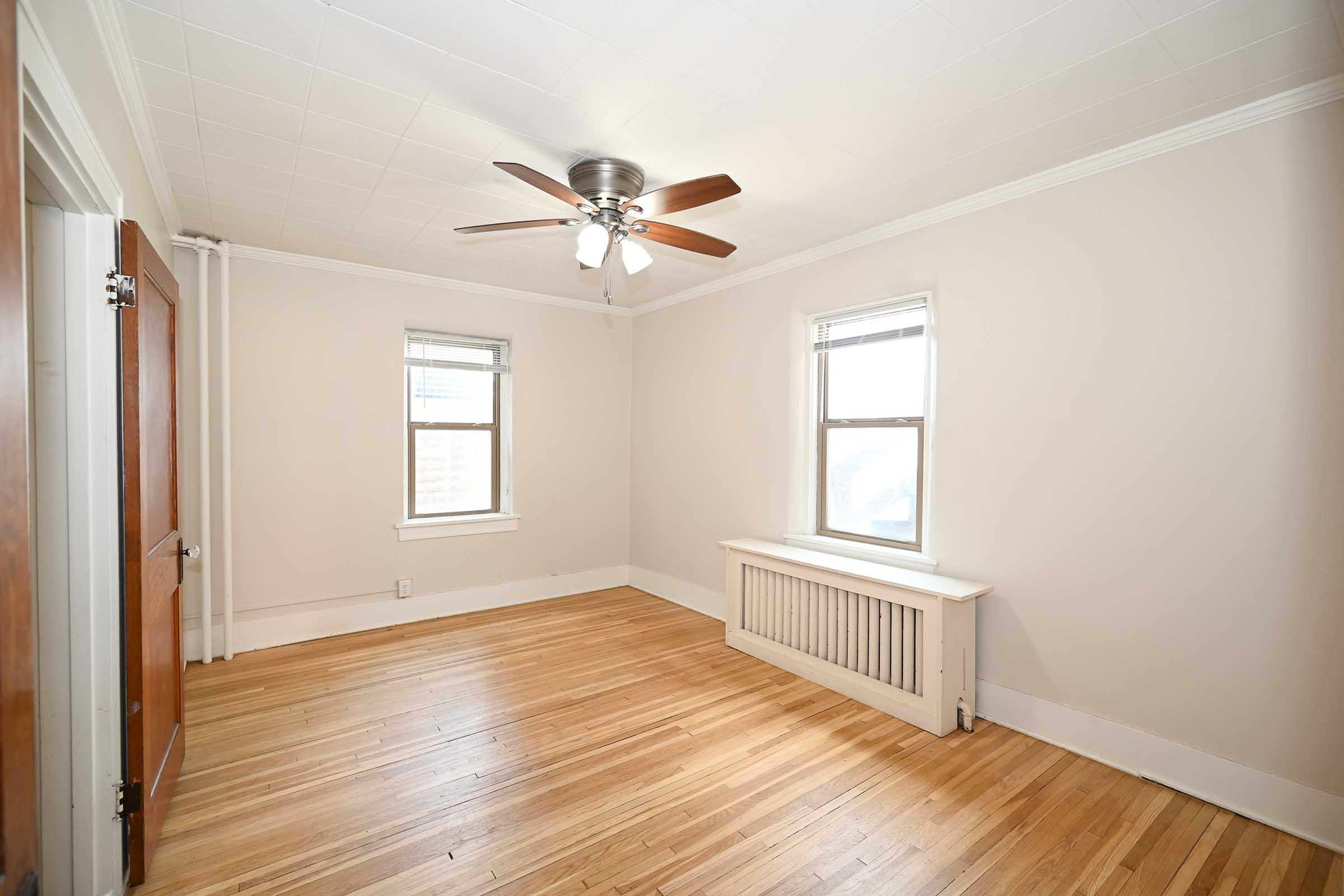 A bright, empty room with light-colored walls and hardwood flooring. Two windows provide natural light, and a ceiling fan is installed. A baseboard heater is located beneath one of the windows, and there are two wooden doors on the left side of the image.