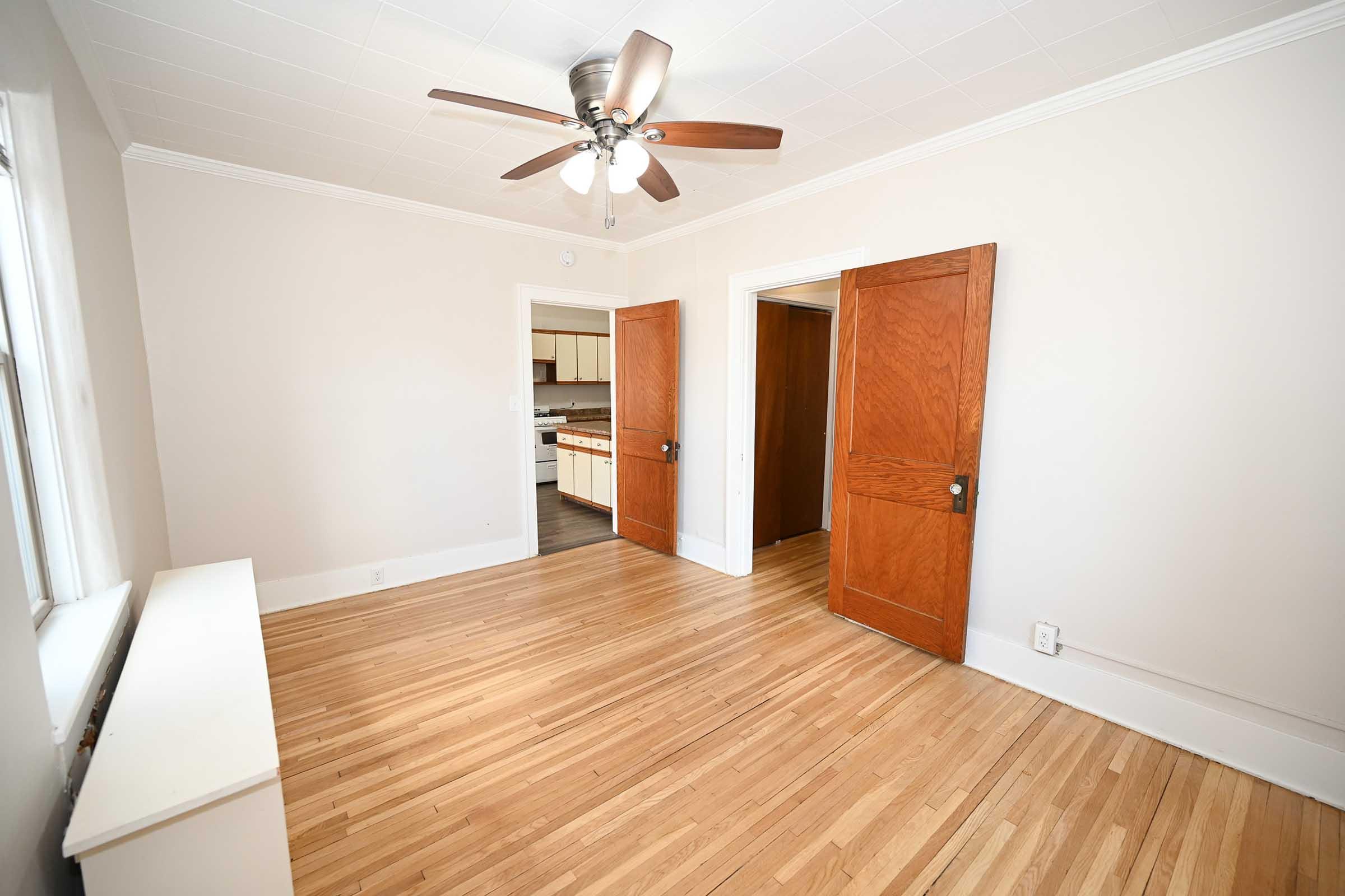A bright and empty room with light-colored walls and hardwood flooring. There are two open wooden doors leading to adjoining spaces. A ceiling fan is installed above, and a white heating unit is visible along one wall. Natural light flows in through the windows, creating an inviting atmosphere.