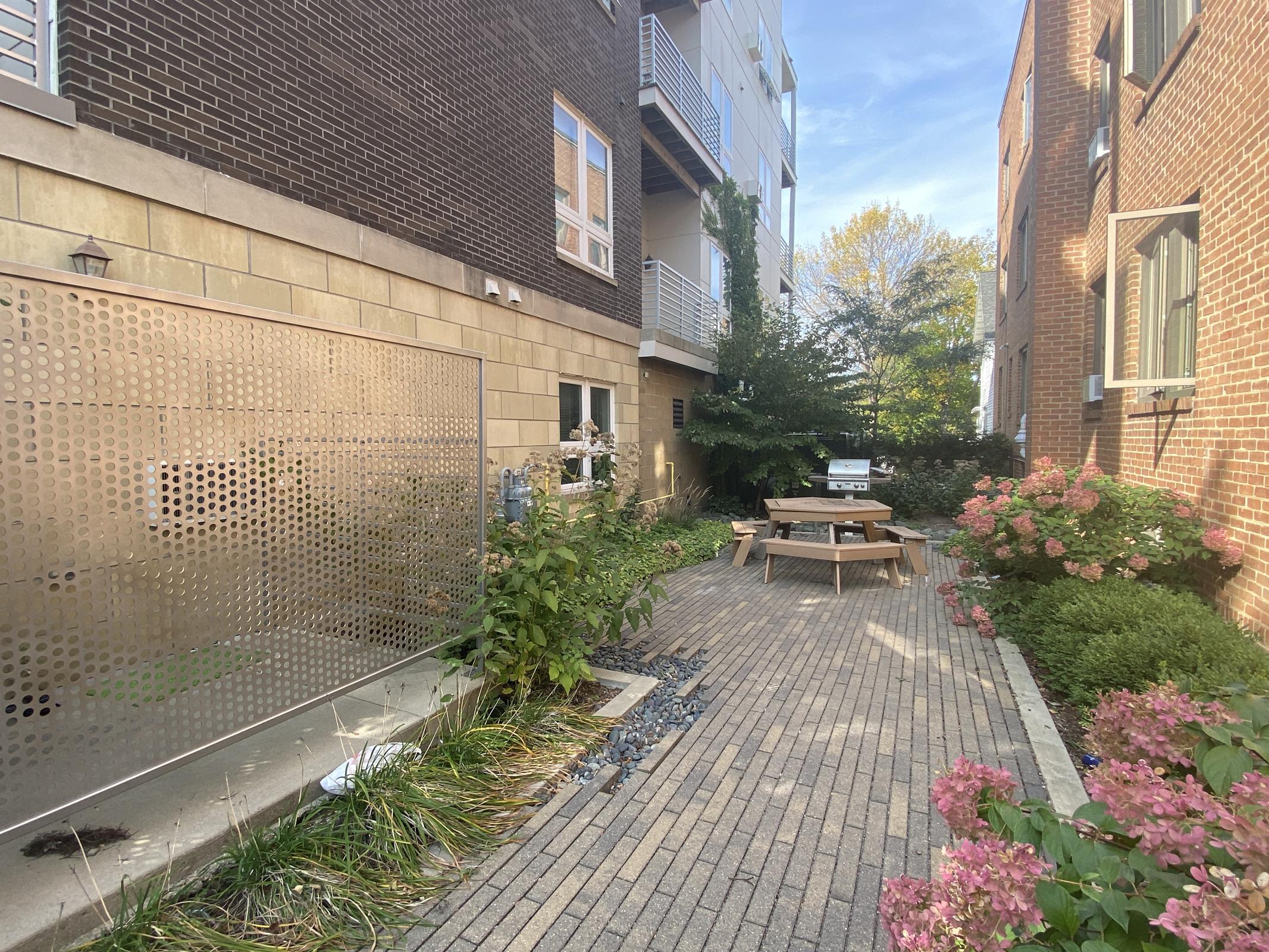 A narrow outdoor pathway surrounded by buildings, featuring a wooden table and benches in the center. Lush green plants and pink flowering shrubs line the path, while a charcoal grill sits nearby. The sky is clear with a hint of sunlight, creating a welcoming atmosphere.