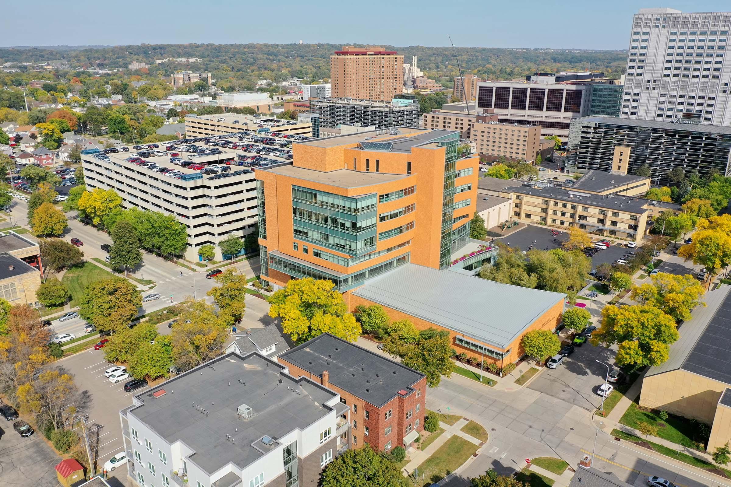Aerial view of a modern multi-story building surrounded by trees and several parked cars, with a mix of urban structures in the background including parking garages and residential buildings. The scene is set in an urban area with a blend of greenery and cityscape.