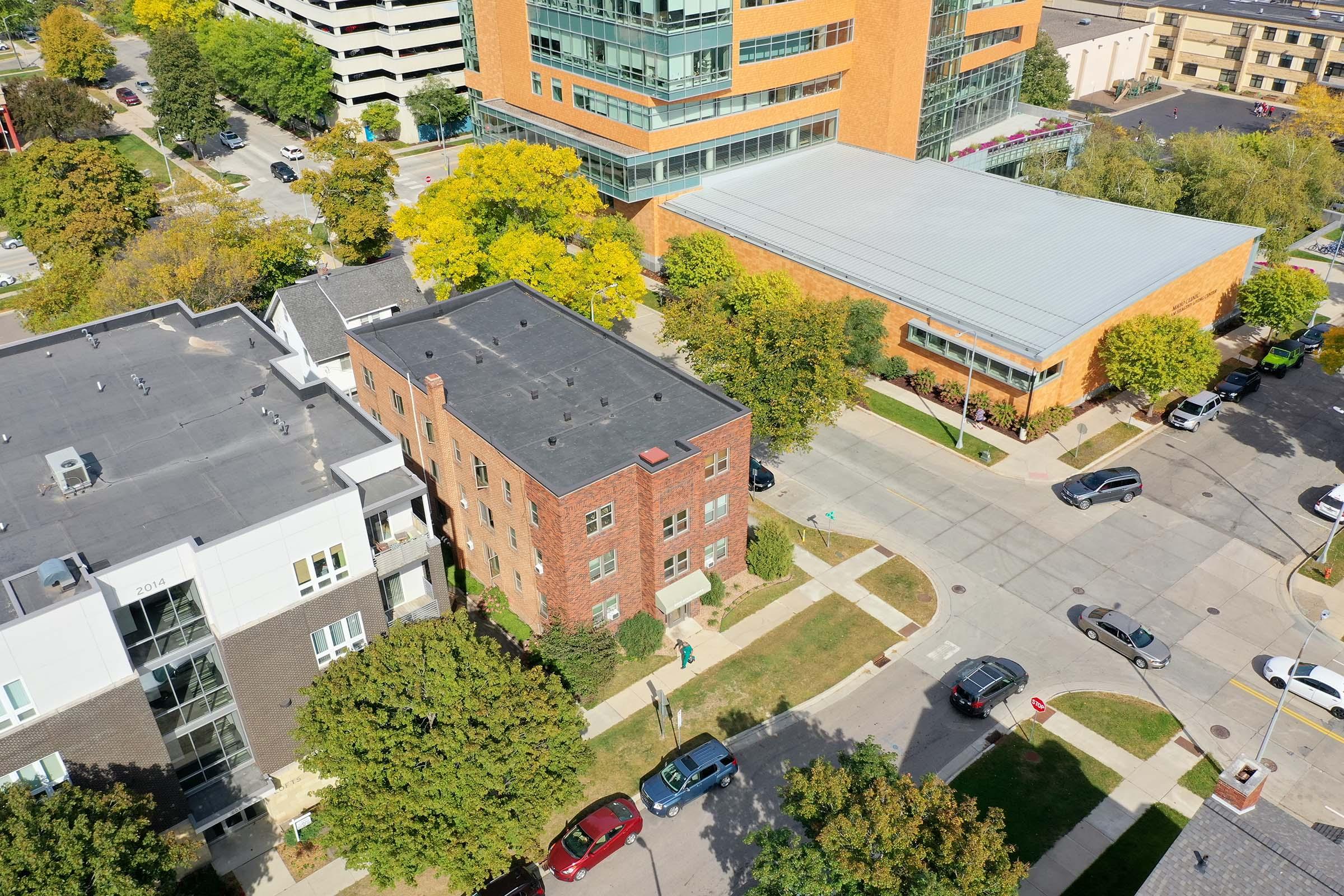 Aerial view of a suburban street featuring a mix of residential buildings and commercial structures. The foreground includes a brick apartment building, while the background shows a modern glass and brick commercial building. Trees with autumn foliage line the streets, creating a vibrant neighborhood scene.