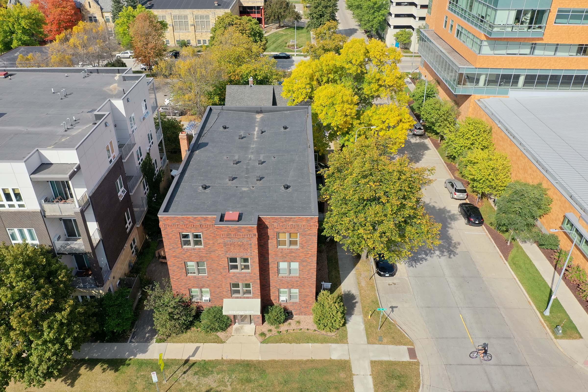 Aerial view of a residential area featuring a brick apartment building with large windows surrounded by greenery. Trees with autumn foliage are visible, and a cyclist rides along the street. Nearby structures include modern buildings and parked cars.