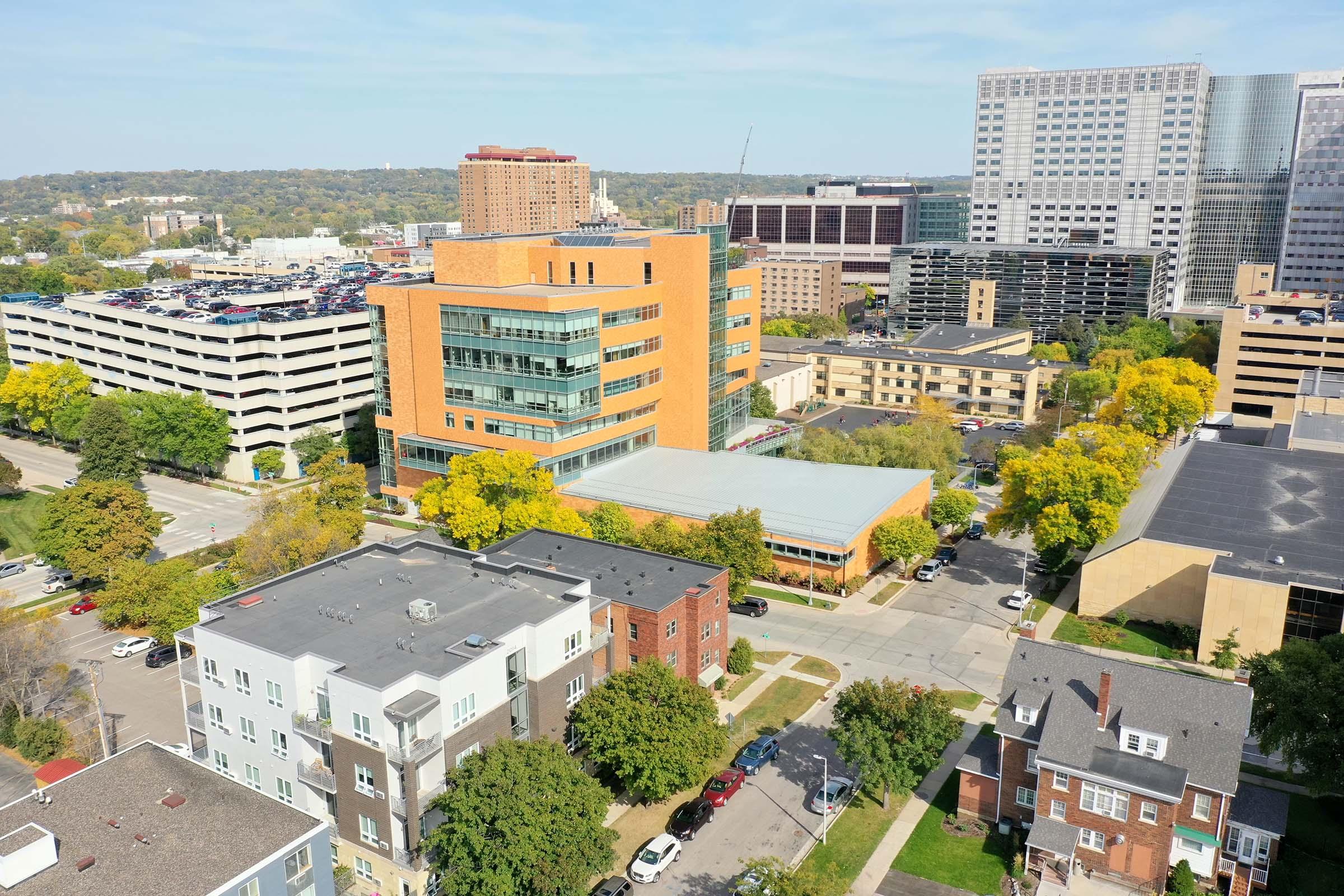 Aerial view of a city block featuring a mix of modern and traditional buildings, including a bright orange structure with glass windows. Surrounding trees are showing autumn colors, and there is a parking garage nearby. In the background, high-rise buildings and an urban skyline complete the scene.