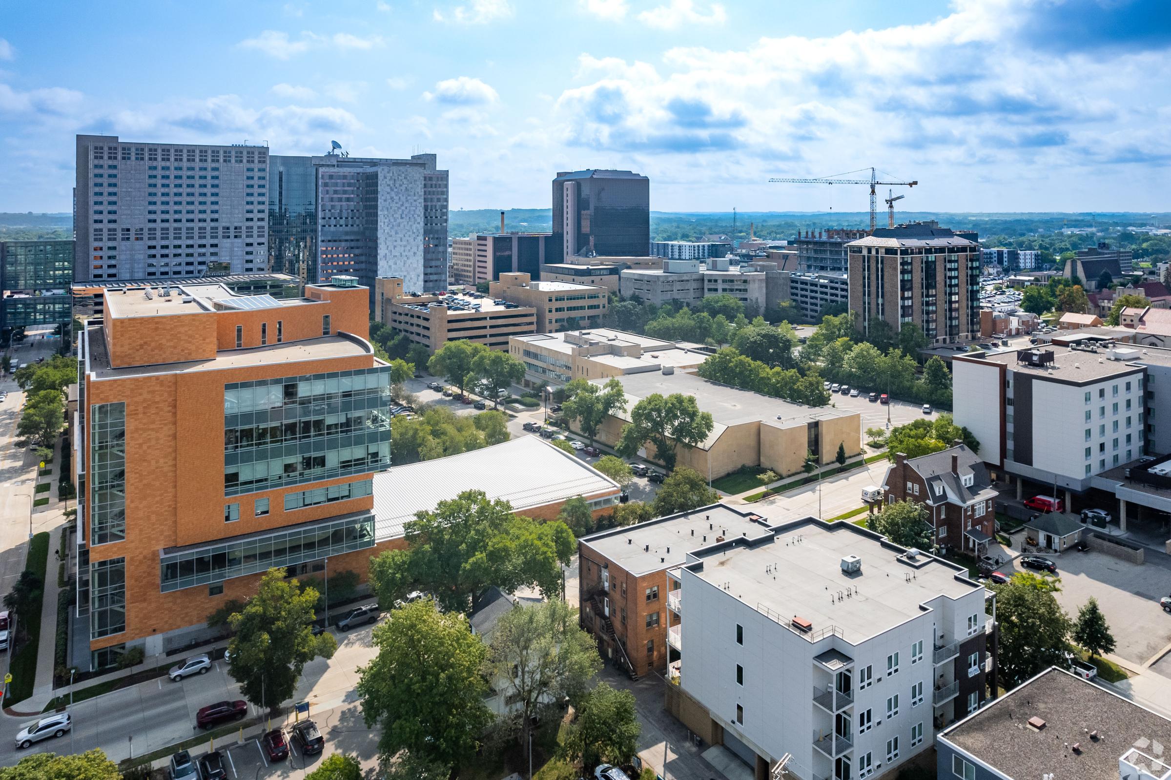 Aerial view of a cityscape featuring a mix of modern buildings, commercial areas, and greenery. The skyline includes tall office structures and residential buildings, along with clear blue skies and scattered clouds. The urban layout shows streets and trees interspersed among the architecture.