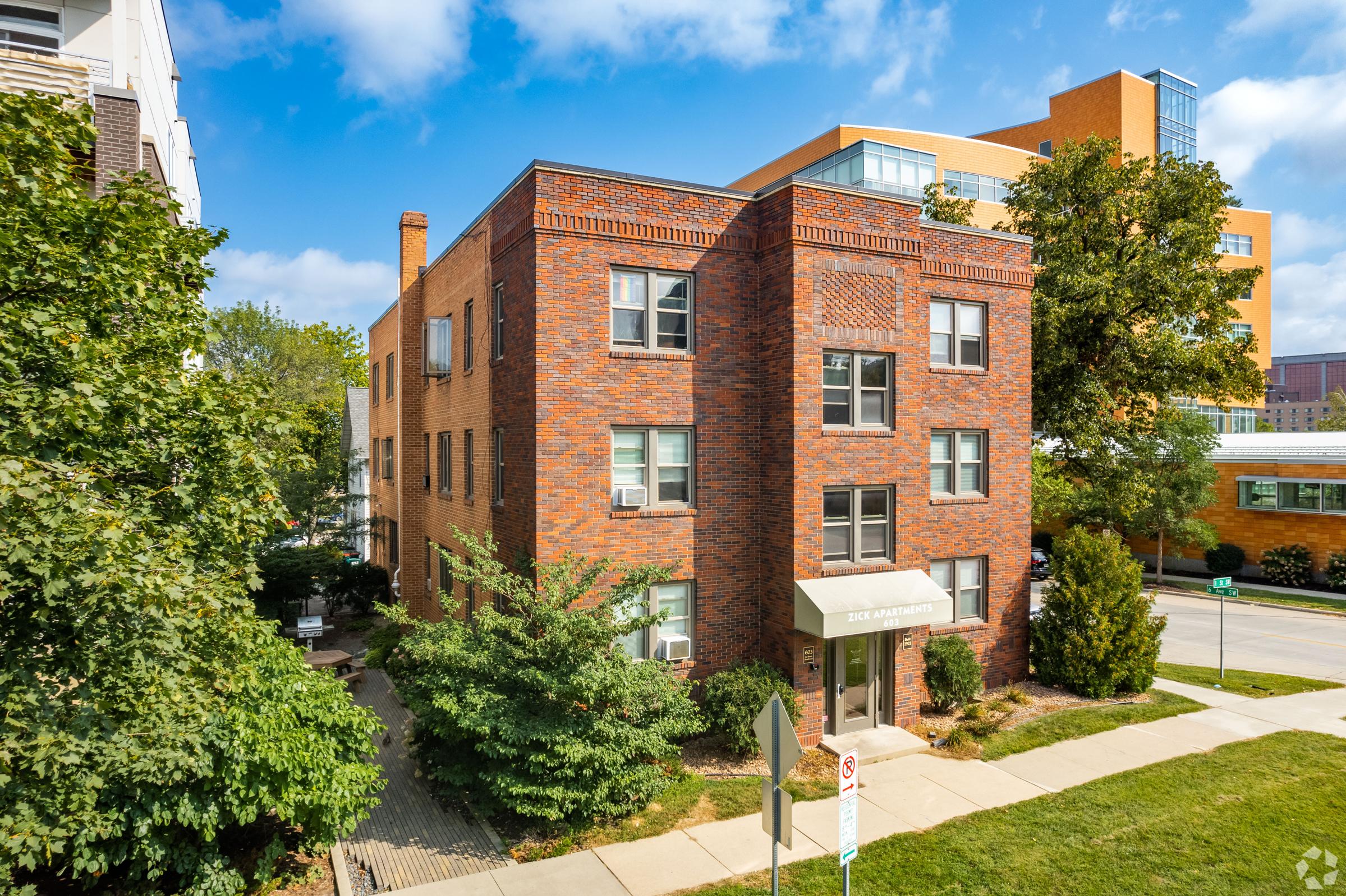 A three-story brick building surrounded by trees and landscaping. The building features large windows and a welcoming entrance with an awning. The environment is sunny with a clear blue sky and additional residential buildings visible in the background. Pathways lead to the entrance and around the property.