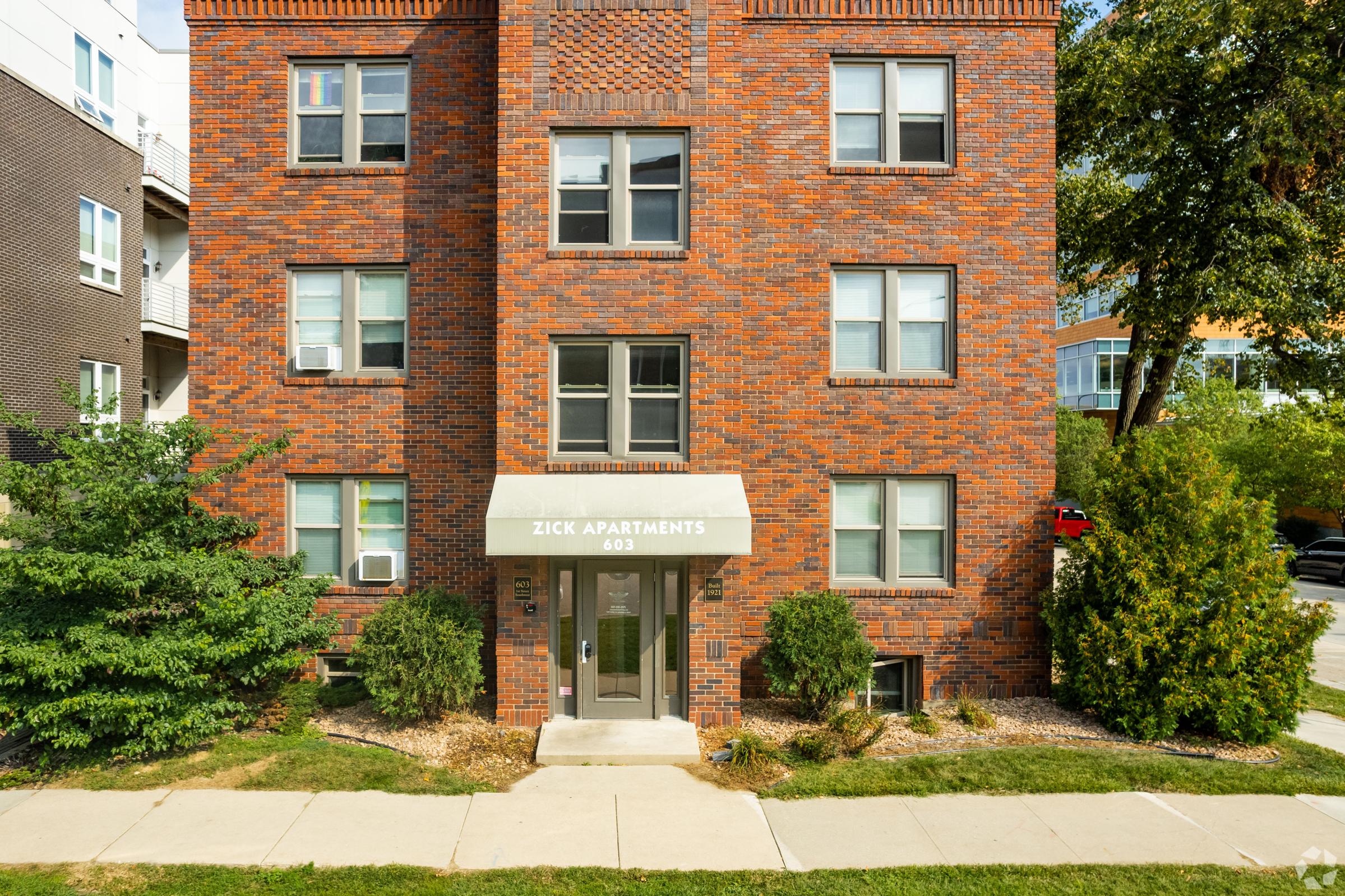 Front view of a brick apartment building labeled "Zick Apartments" with a canopy above the entrance. The building features multiple windows and is surrounded by greenery, including shrubs. A sidewalk leads up to the entrance, and there are other residential buildings visible in the background.