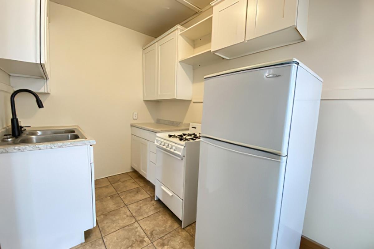 A small, modern kitchen featuring white cabinetry, a stainless steel sink, a white stove, and a refrigerator. The floor is tiled, and natural light enters through the window, creating a bright and clean atmosphere. The kitchen is functional with a simple layout, ideal for small spaces.
