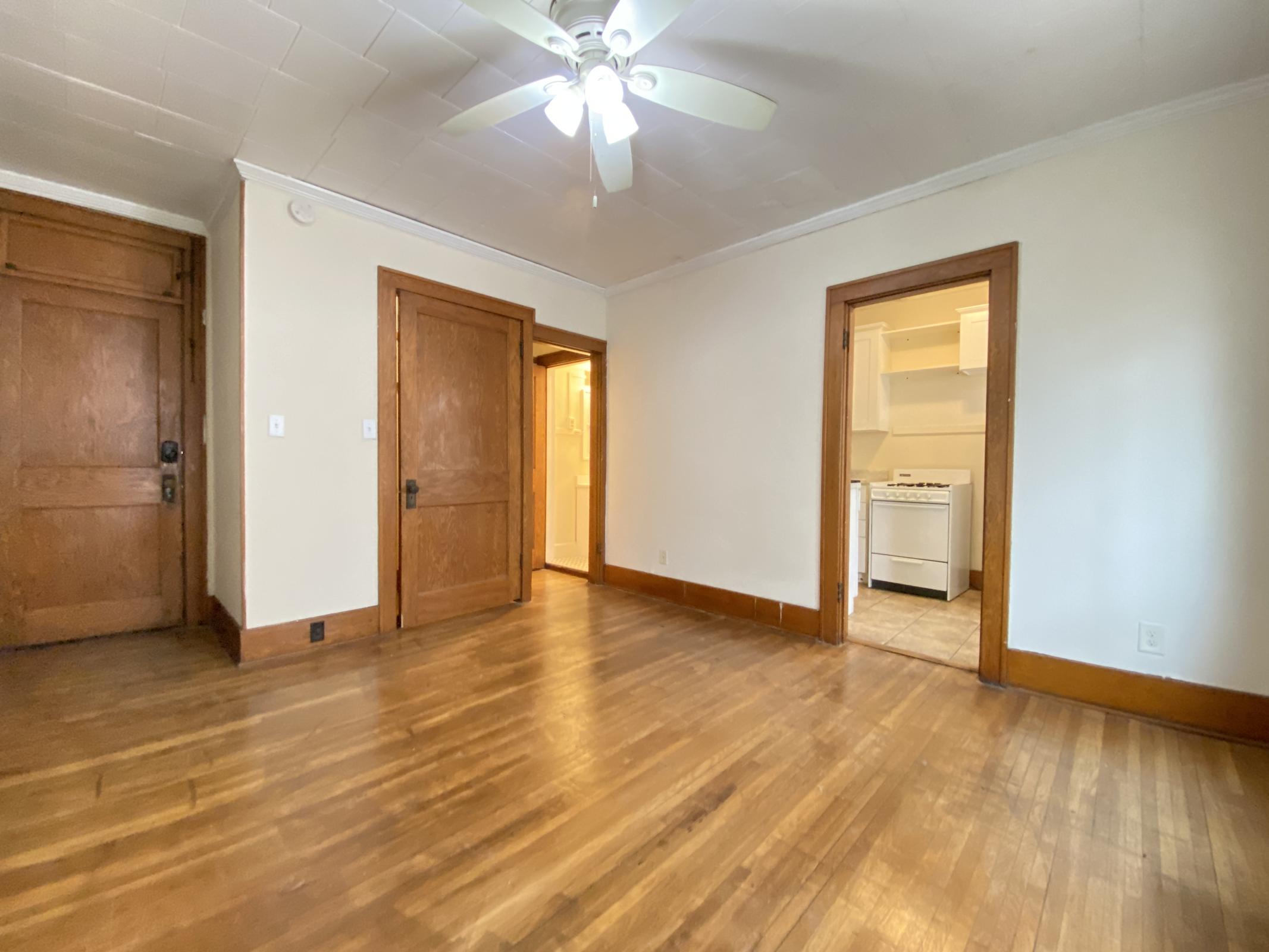 A spacious, empty room with hardwood floors, a ceiling fan, and two wooden doors leading to adjacent areas. The room features light-colored walls and a neutral color scheme, creating a bright and inviting atmosphere. The doorway on the right opens into a kitchen area.