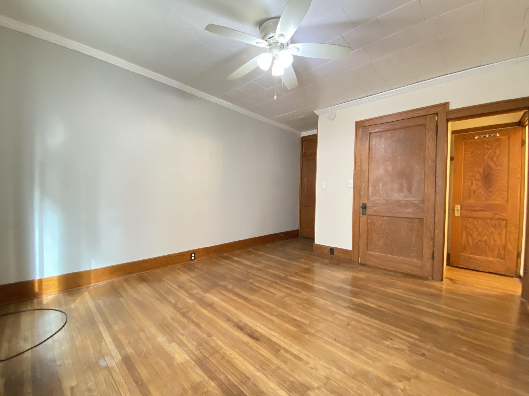 A spacious, empty room featuring polished hardwood floors, a ceiling fan, and light-colored walls. One corner has a doorway leading to a wooden door, while there are no visible furnishings, creating an open and airy atmosphere. Natural light enters from a window on the left.