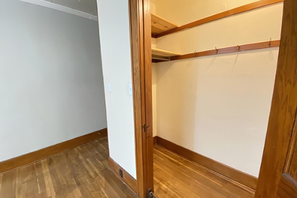 Interior view of a small room with a closet. The closet features a wooden rod for hanging clothes and hooks on the wall. The floor is hardwood, and the walls are painted in light colors. A doorway leads into the room from the left, showcasing a minimalist design and natural light.