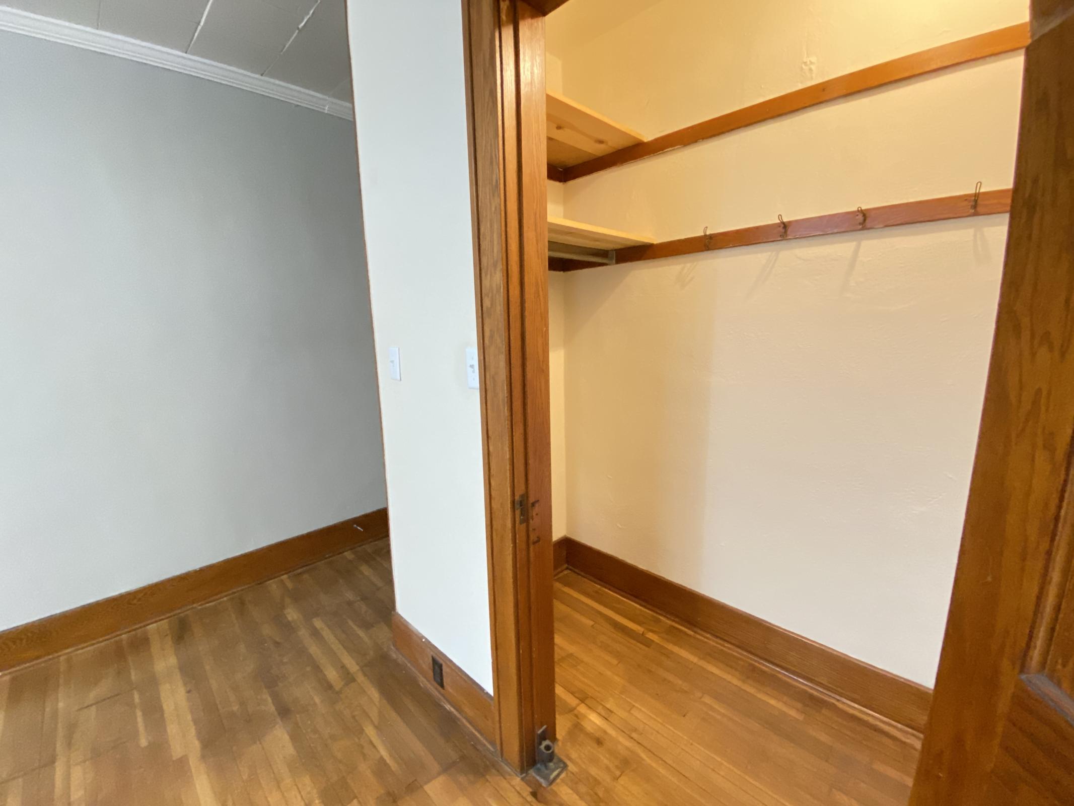 A view of a small, empty closet with wooden shelving and hooks, adjacent to a room with wooden flooring and neutral-colored walls. The doorway leading into the closet is open, revealing the minimalist design and lack of furnishings inside.