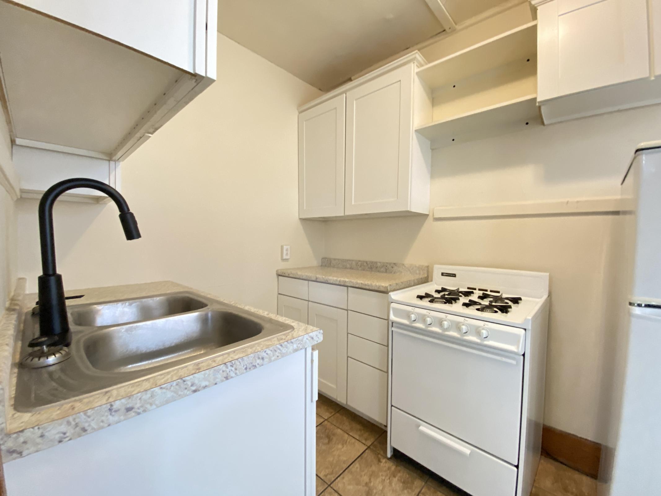 A small kitchen featuring a sink with a black faucet, a white gas stove, and white cabinetry. The countertops are a light-colored granite, and the walls are painted a neutral shade. Shelving above the sink is empty, and the floor has beige tiles.