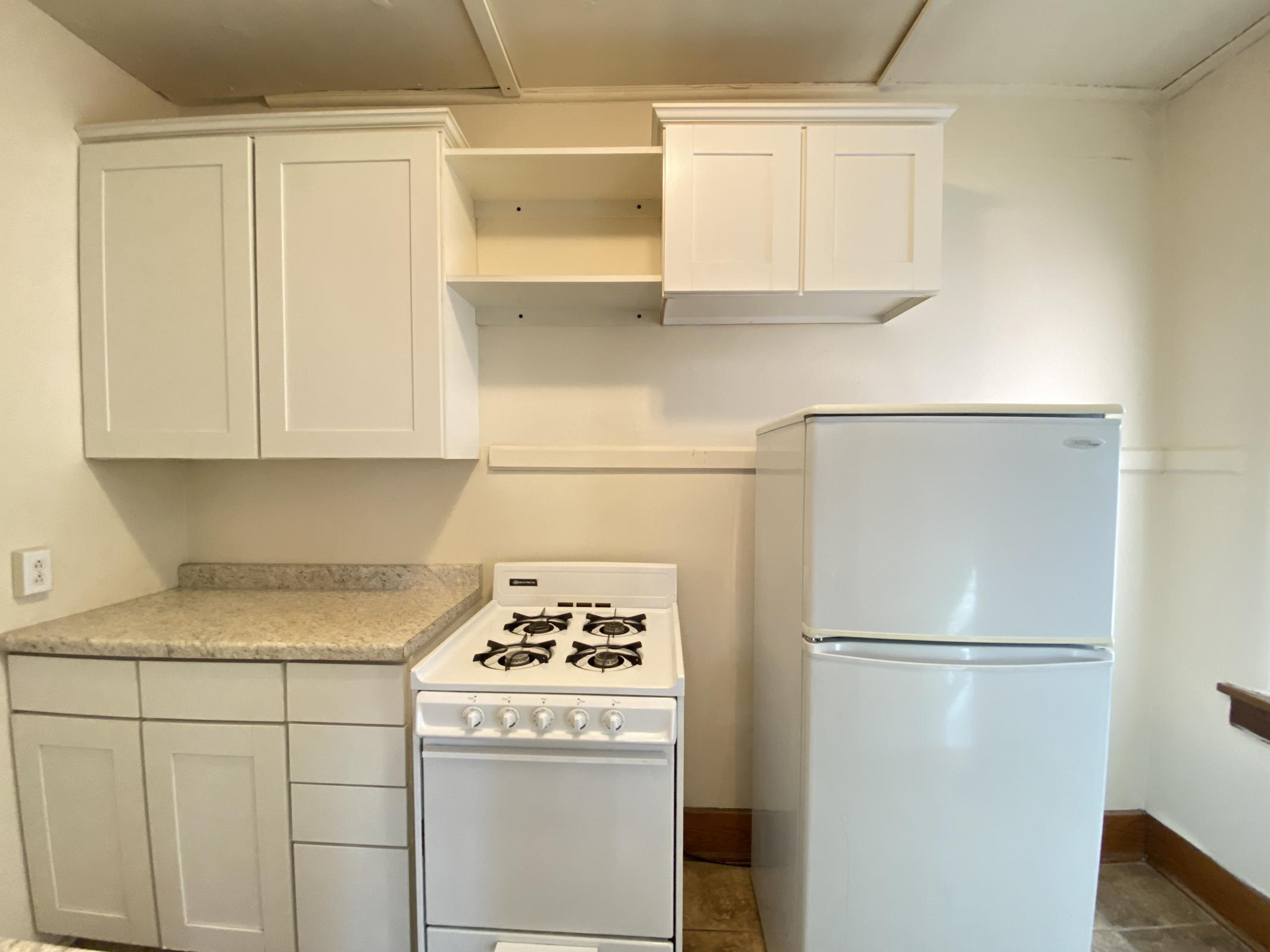 A small kitchen featuring white cabinets, a granite countertop, a white gas stove with four burners, and a white refrigerator. The walls are painted light colors, creating a bright and clean appearance. A few shelves are above the countertop, providing additional storage space.