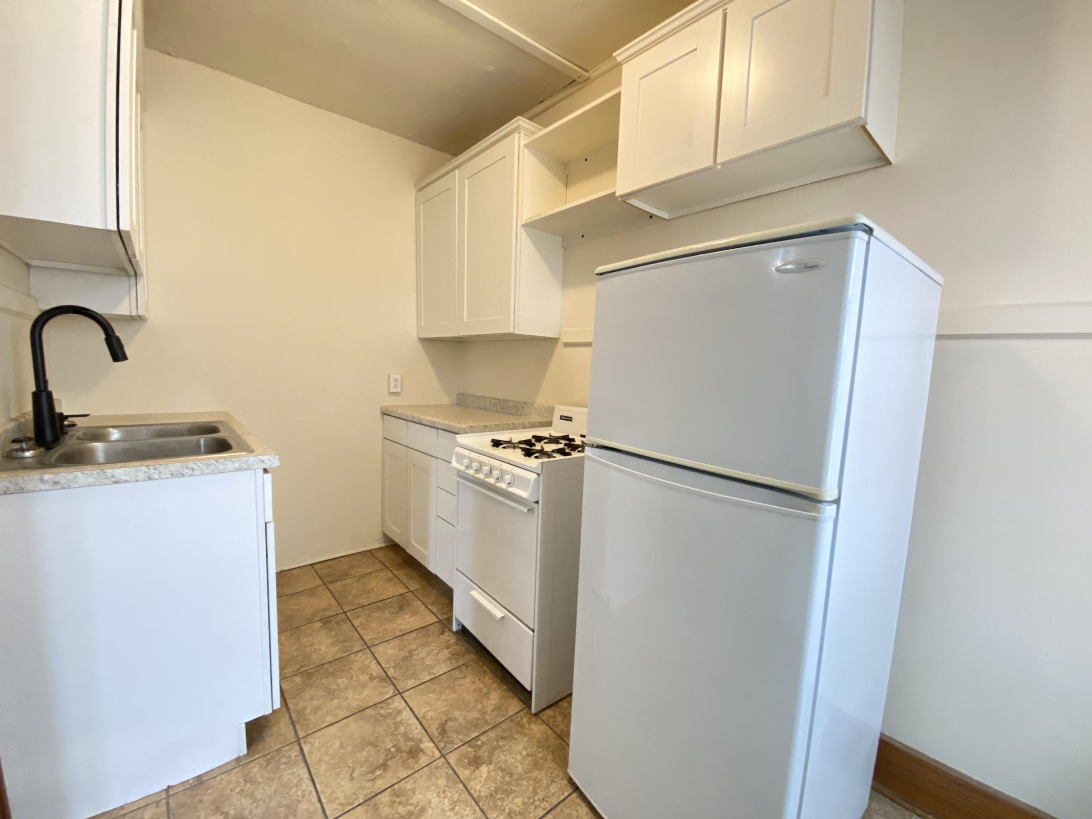 A small kitchen featuring white cabinets, a granite countertop, a stainless steel sink with a black faucet, a white stove, and a white refrigerator. The floor is tiled, and the walls are painted in a light color, creating a bright and clean atmosphere.