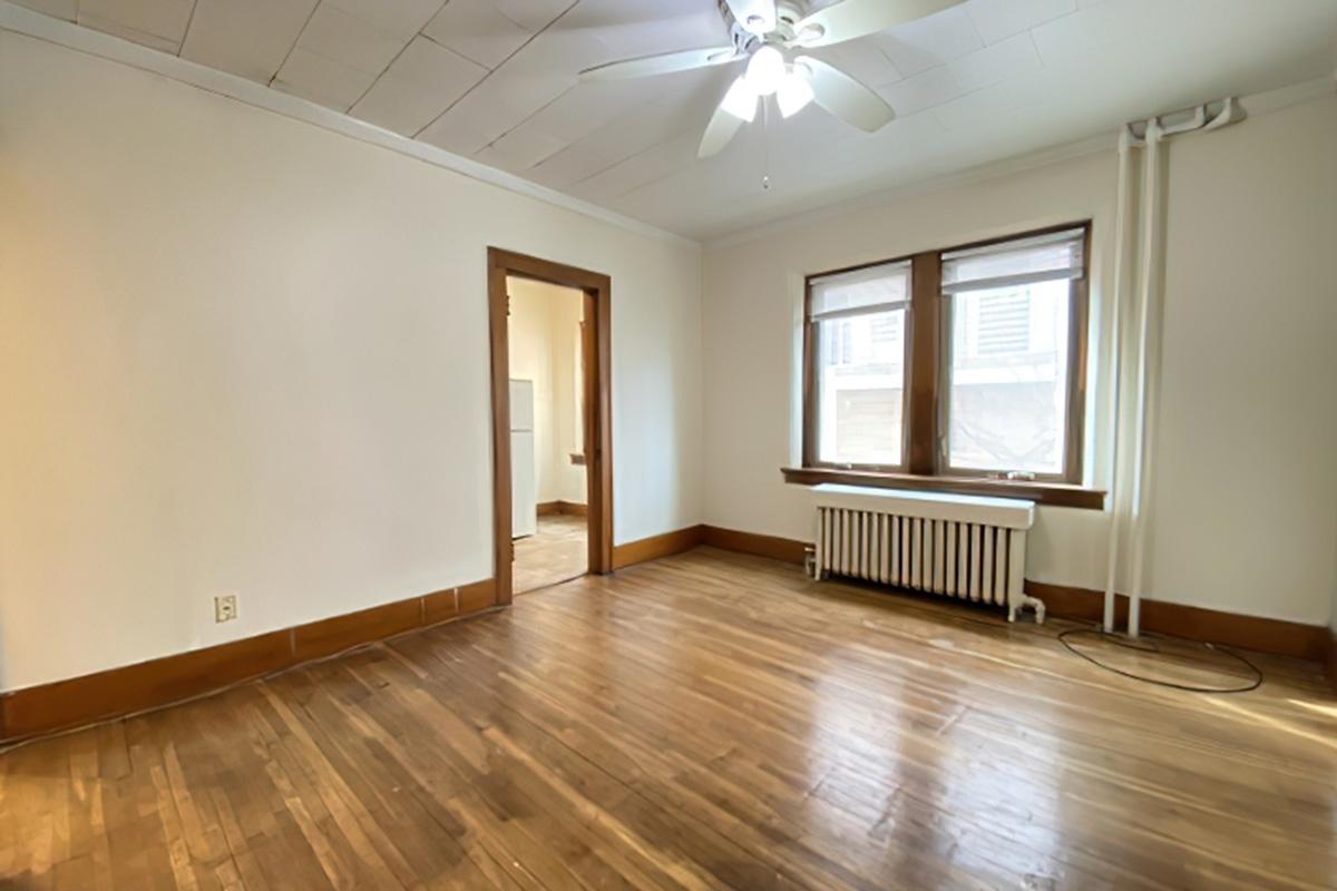 An empty room featuring hardwood floors, a ceiling fan, and a radiator. There are two windows on one side allowing natural light, and a doorway leading to another area. The walls are painted a light color, providing a spacious and bright appearance.