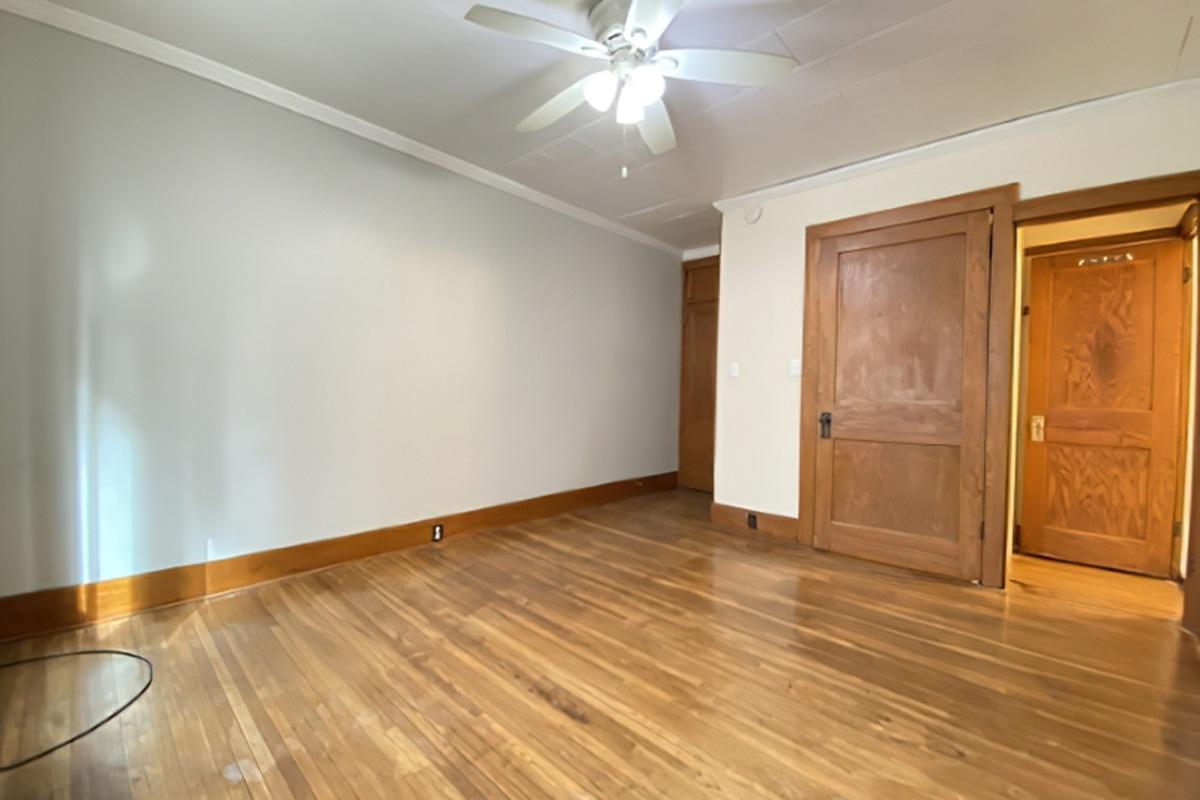 A spacious, empty room with wooden flooring, light gray walls, and a ceiling fan. There are two wooden doors with ornate handles on the right side, one partially open. The room is well-lit, showcasing a clean and minimalist design.
