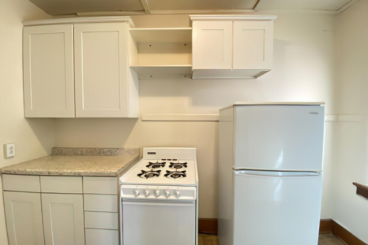 A compact kitchen featuring white cabinets, a stove with four burners, and a refrigerator. The cabinets are above a countertop, and there's an empty shelf for additional storage. The overall design is simple and functional, suitable for small living spaces.