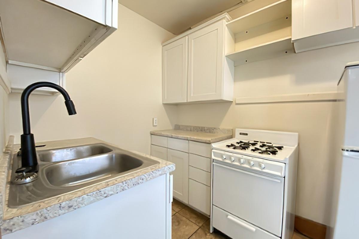 A small, modern kitchen featuring a stainless steel sink with a black faucet, white cabinets, a white gas stove, and beige tile flooring. The cabinetry is simple and the space is well-lit, providing a clean and functional cooking area.