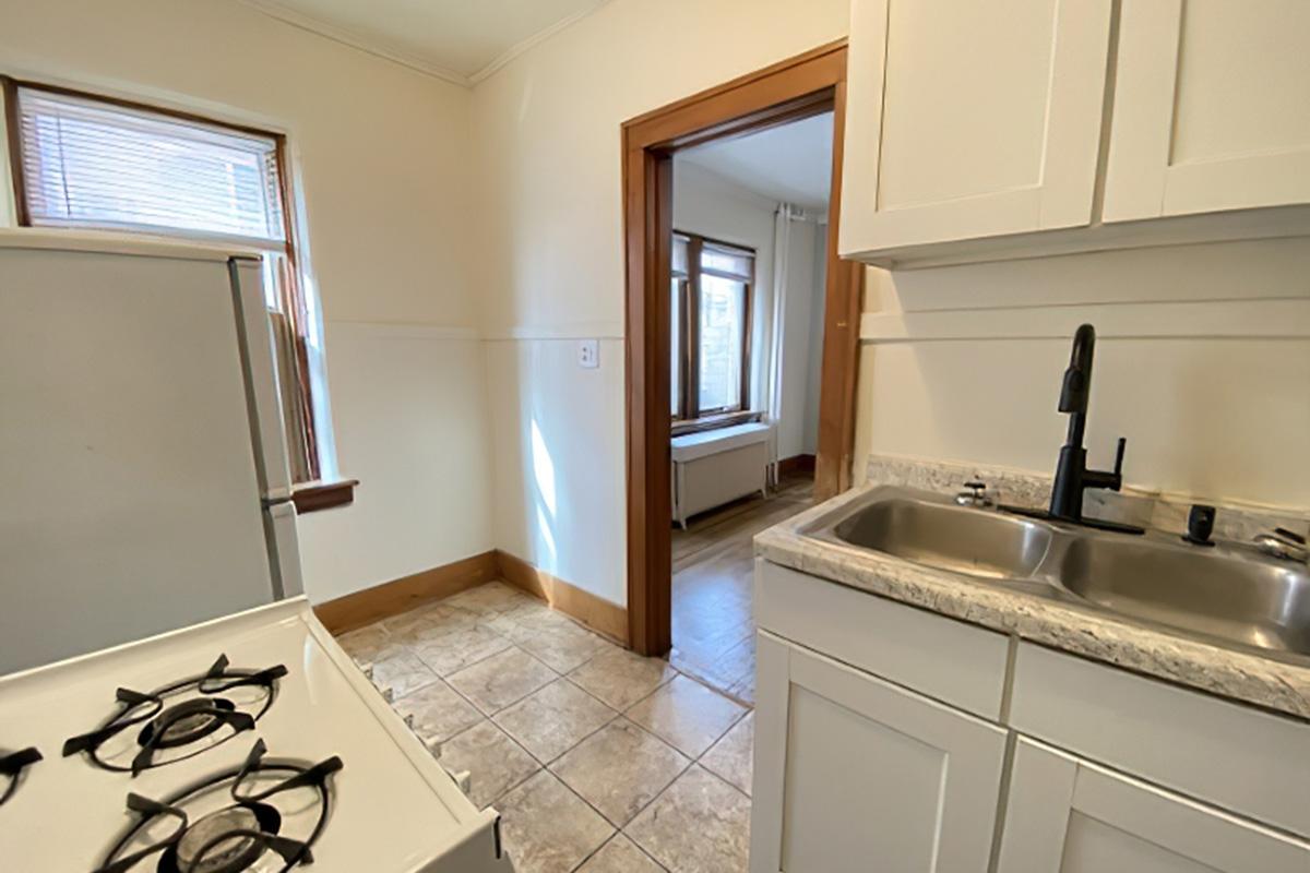 A small kitchen featuring a white gas stove, sink with a black faucet, and light-colored cabinetry. The space has tiled flooring and a window providing natural light. A doorway leads to another room with wooden flooring and large windows, adding to the homey atmosphere.