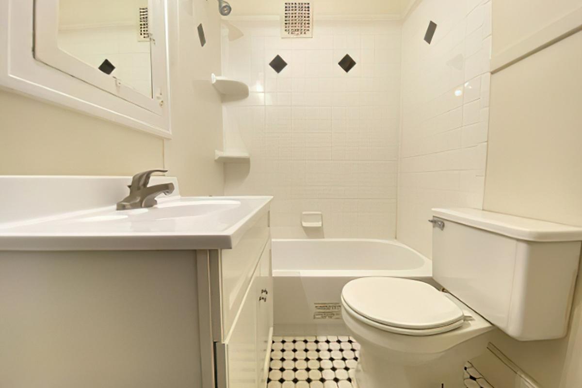 A clean, well-lit bathroom featuring a white sink with a modern faucet, a bathtub with a shower, and a toilet. The walls are tiled in white with black accents, and the floor has a patterned black and white tile design. Shelves are mounted on the wall for storage.