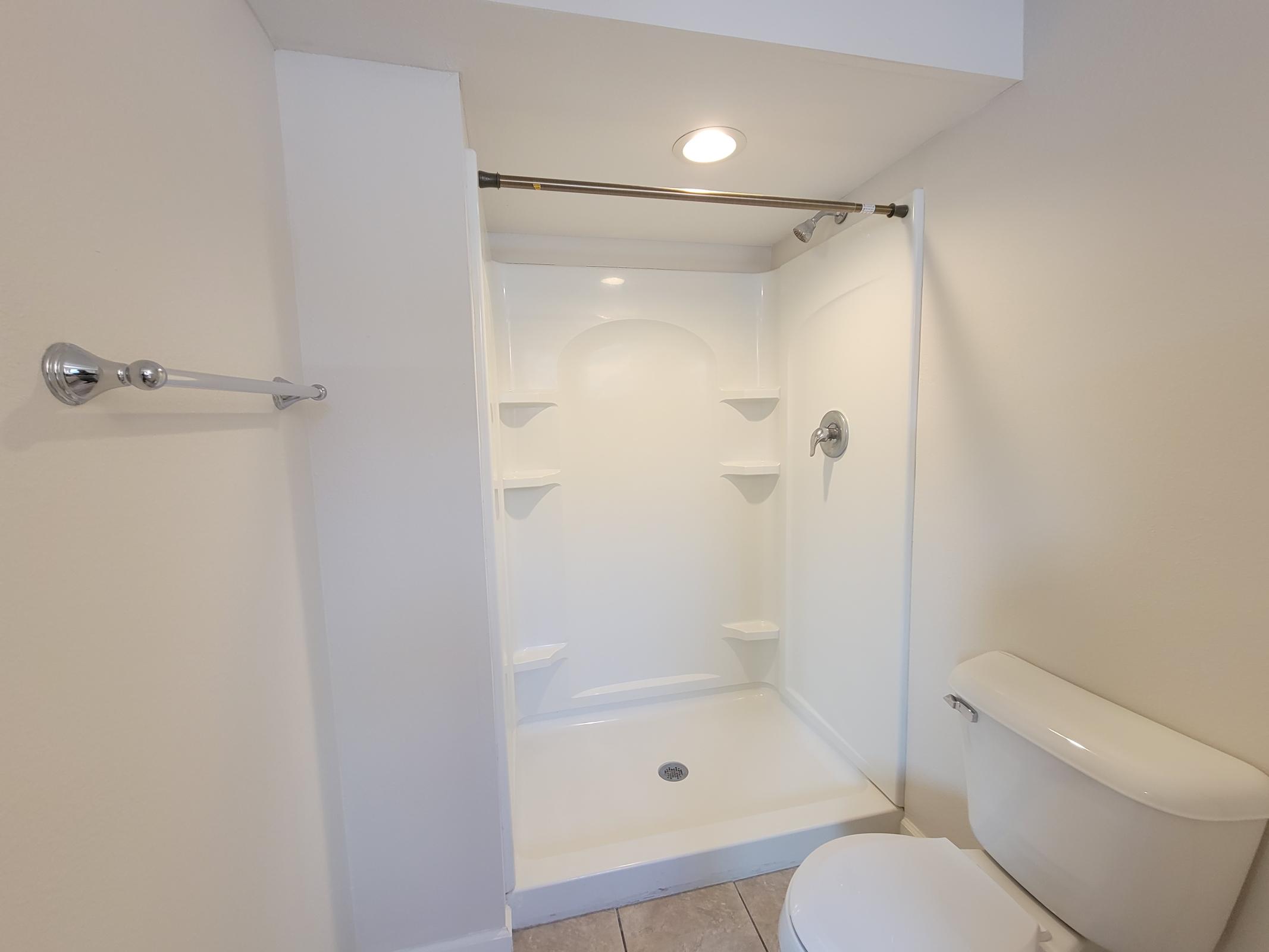A clean and modern bathroom featuring a white shower enclosure with built-in shelves, a chrome showerhead, and a white toilet. The walls are plain and light-colored, and there is a towel bar mounted on the wall. The floor has beige tiles, creating a simple, bright aesthetic.