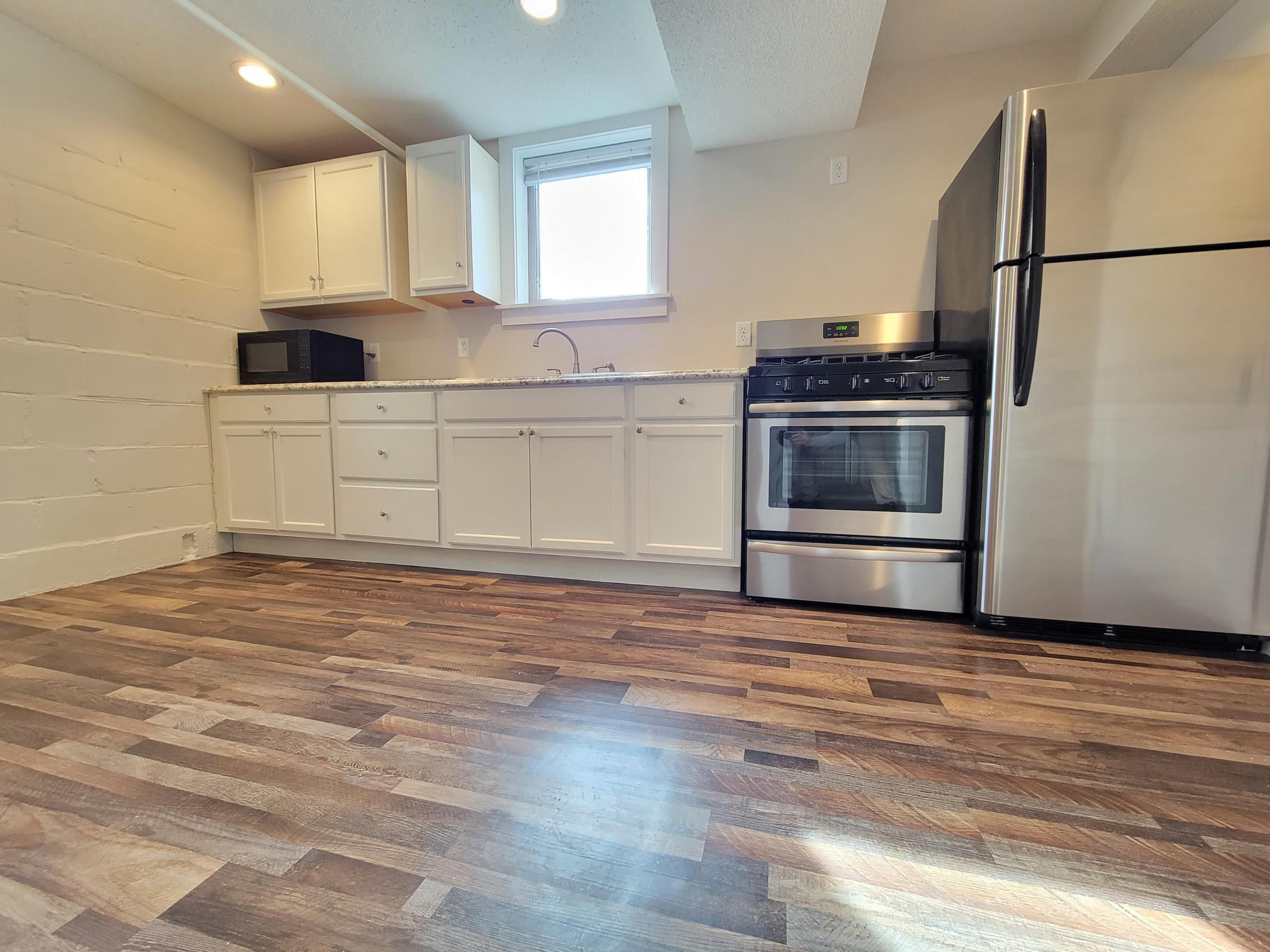 A modern kitchen featuring white cabinets, a stainless steel oven and refrigerator, a microwave, and a sink. The countertop is made of granite, and the flooring is wooden. Natural light enters through a window, creating a bright and inviting atmosphere.
