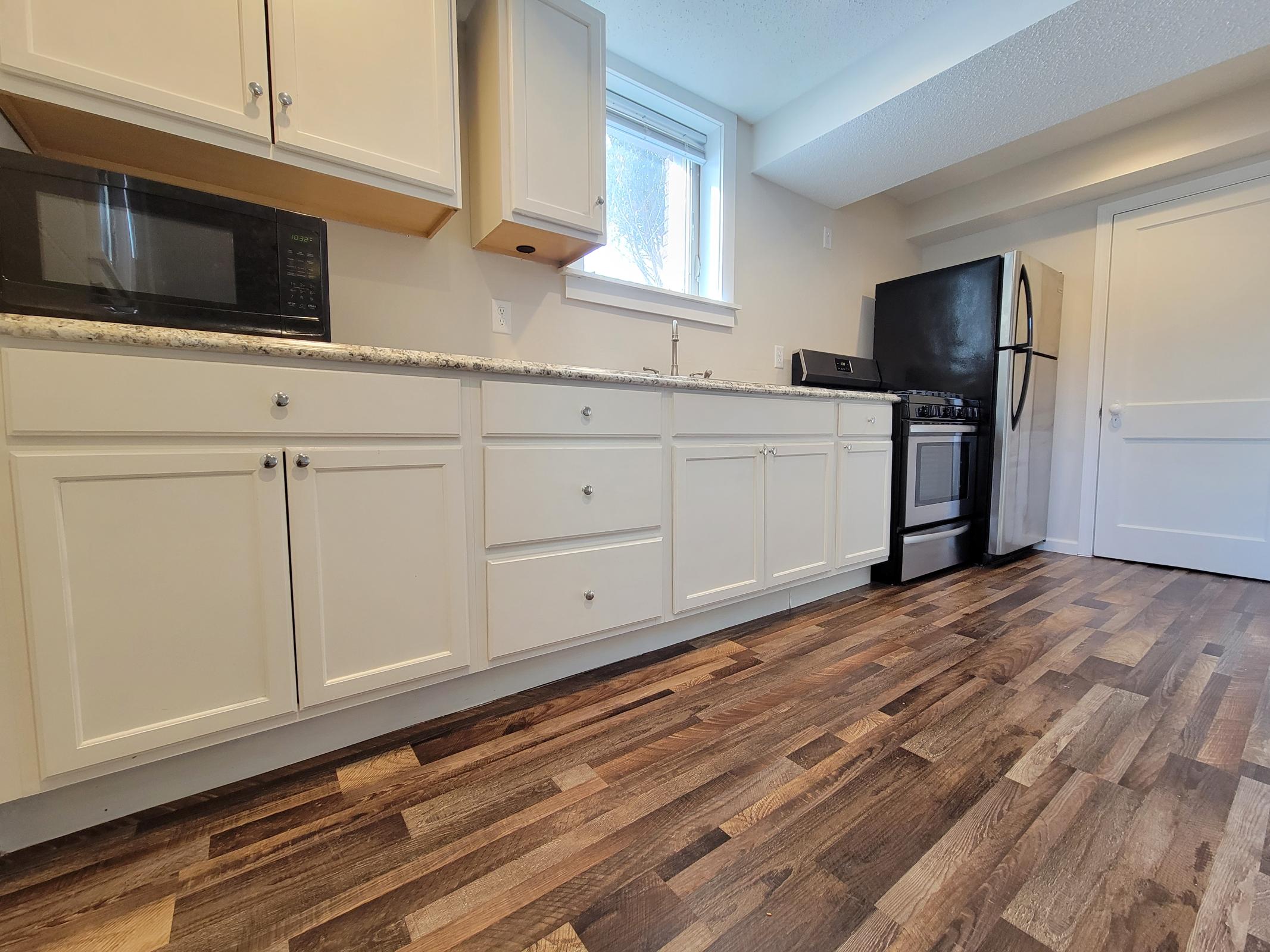 A modern kitchen featuring white cabinetry, a microwave, and a black refrigerator beside a stainless steel stove. The countertop is light-colored with a subtle pattern, and the floor is made of dark wood planks. Natural light enters through a window, illuminating the space.