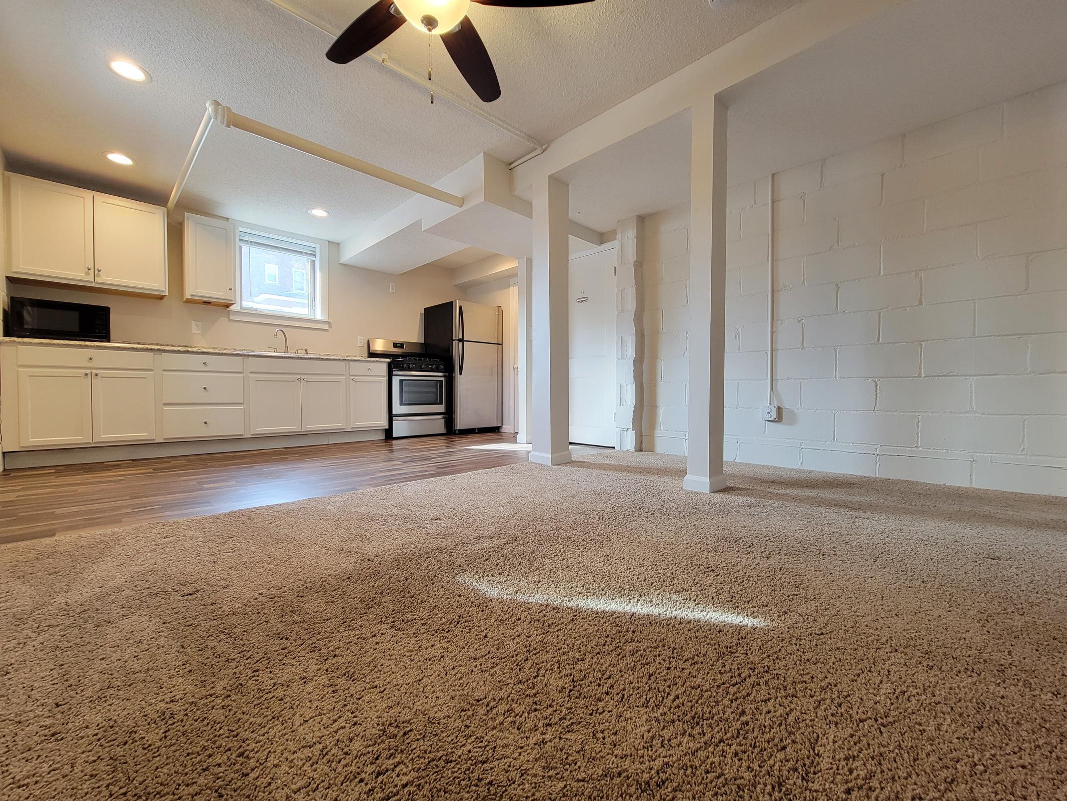 A bright, spacious basement apartment features a kitchen with white cabinets, a microwave, and a stainless steel oven. The living area has carpeted flooring and a ceiling fan, with white painted walls and natural light coming from a window. The layout includes a small kitchenette and open space.