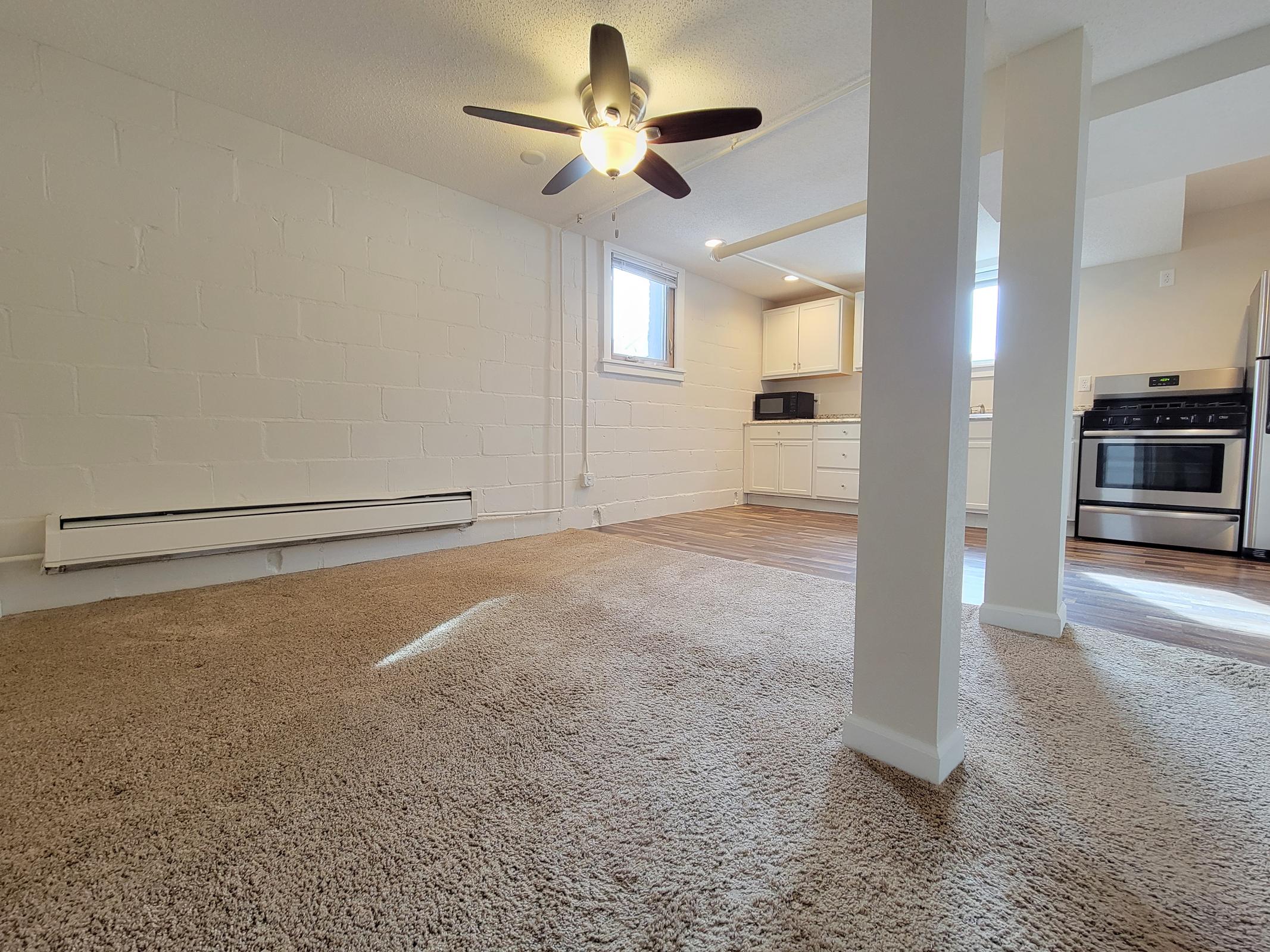 A bright, spacious room with a ceiling fan and beige carpet. On one side, there is a kitchenette featuring white cabinets, a microwave, and a stainless steel stove. A window allows natural light to enter, and the walls are painted white, providing a clean and modern look.
