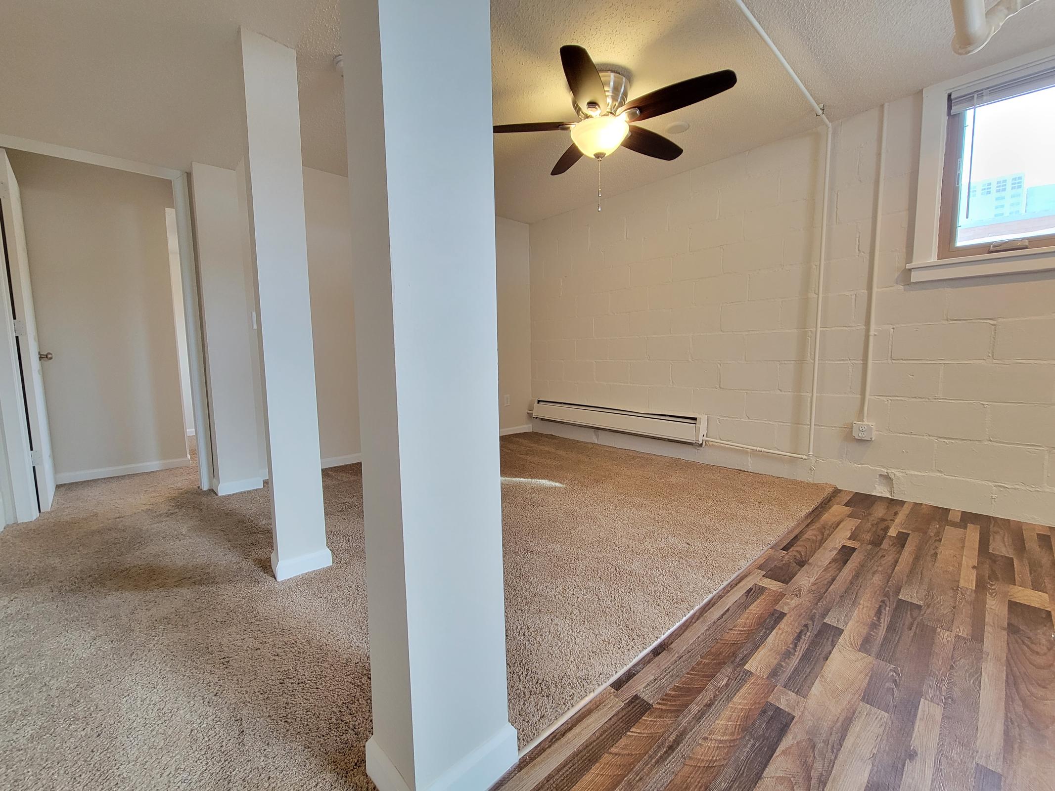A spacious, well-lit room with a ceiling fan. The flooring features a mix of carpet and hardwood. There are two visible column supports, a window allowing natural light, and an open layout ideal for living or recreational use. The walls are painted white, and the setting appears modern and clean.