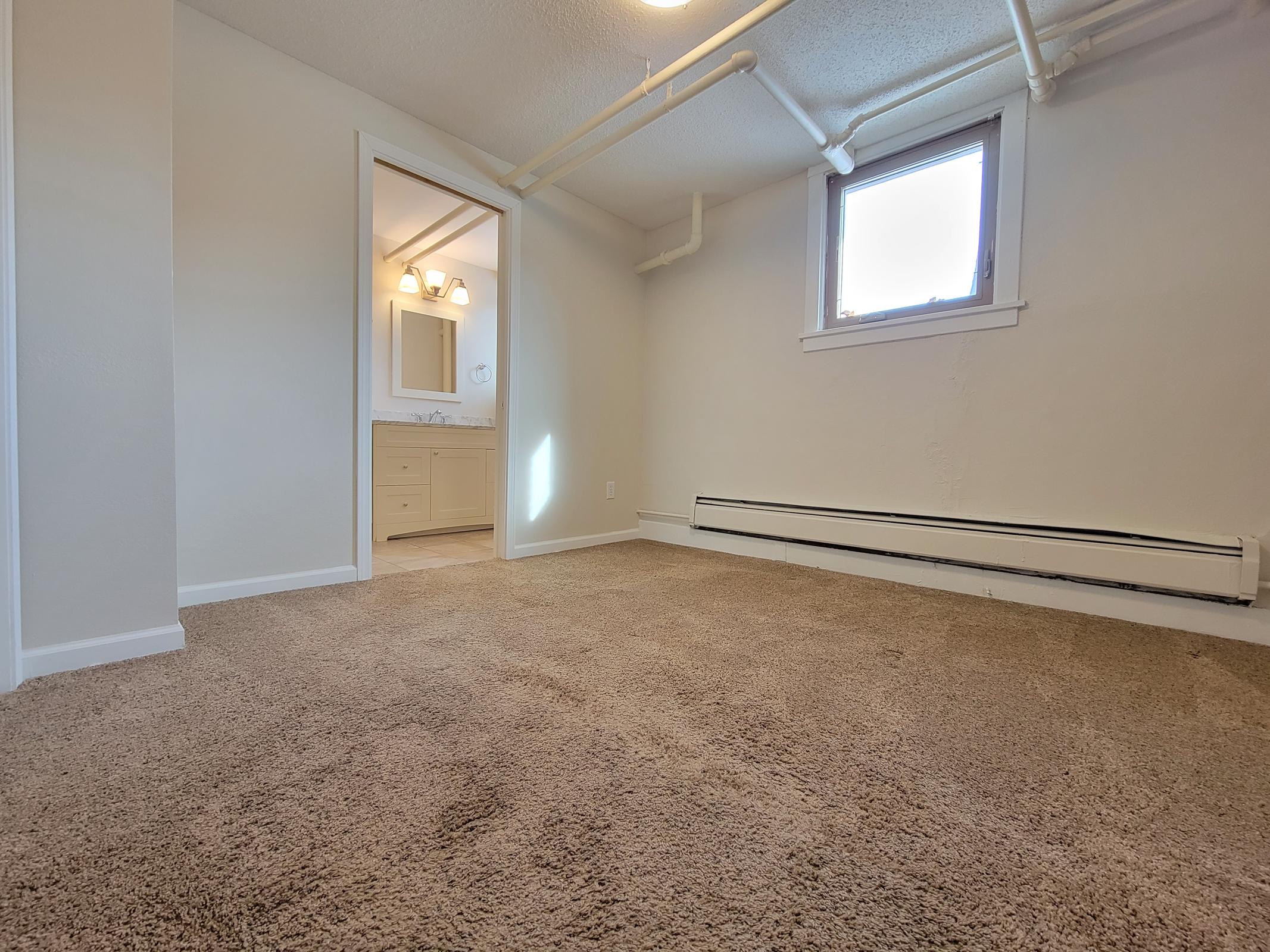 Empty room with beige carpet, featuring a small window on the wall. A doorway leads to a bathroom with vanity lighting visible. The walls are painted white, and there are exposed pipes along the ceiling. The overall atmosphere is bright and clean, suitable for various purposes.