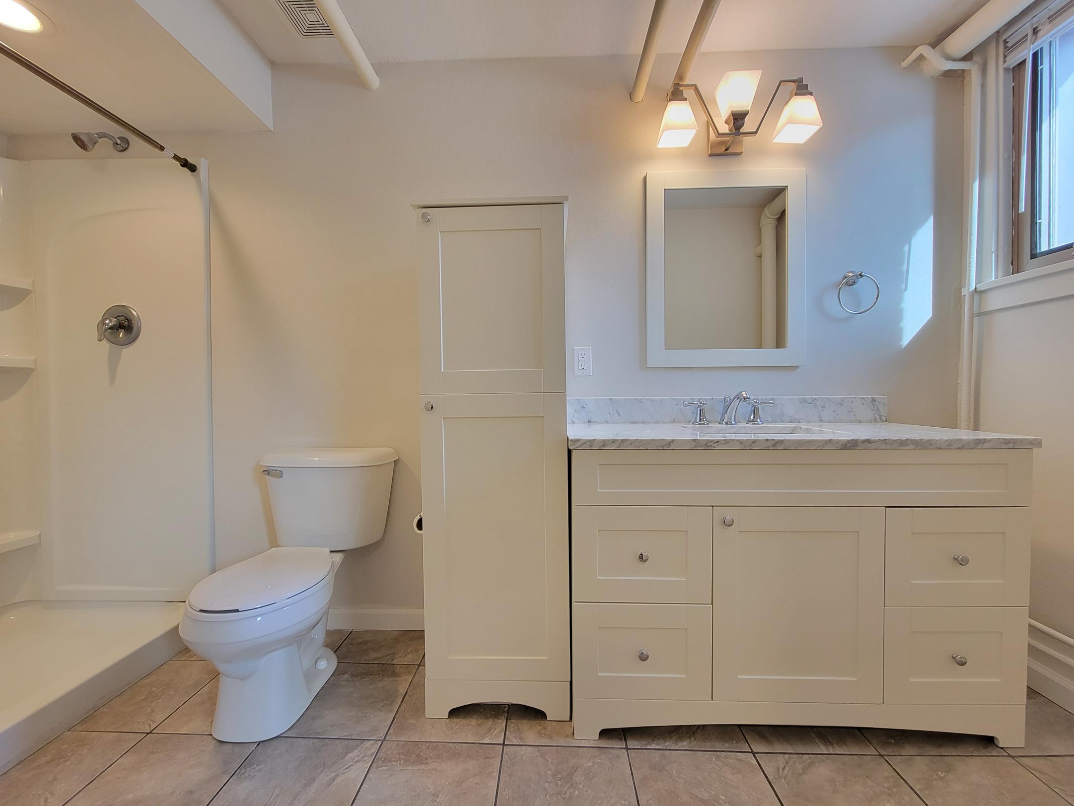 A modern bathroom featuring a white toilet, a shower with a glass door, and a light-colored vanity with a marble countertop and dual sinks. The space is well-lit with natural light and has neutral tile flooring. A mirror is mounted above the sinks, and there is a tall cabinet for storage.