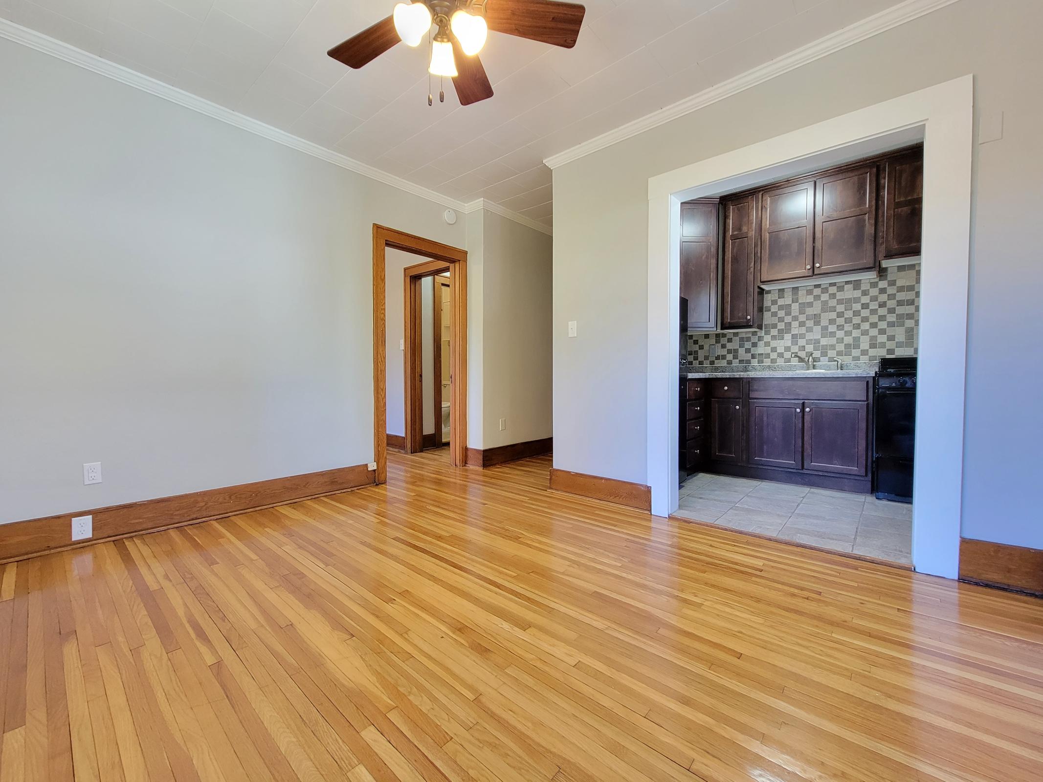 A spacious, well-lit living area featuring hardwood floors, a ceiling fan, and light gray walls. An open doorway leads to a kitchen area with dark wood cabinets and tiled flooring, creating a cozy and inviting atmosphere. The overall design combines modern and classic elements.