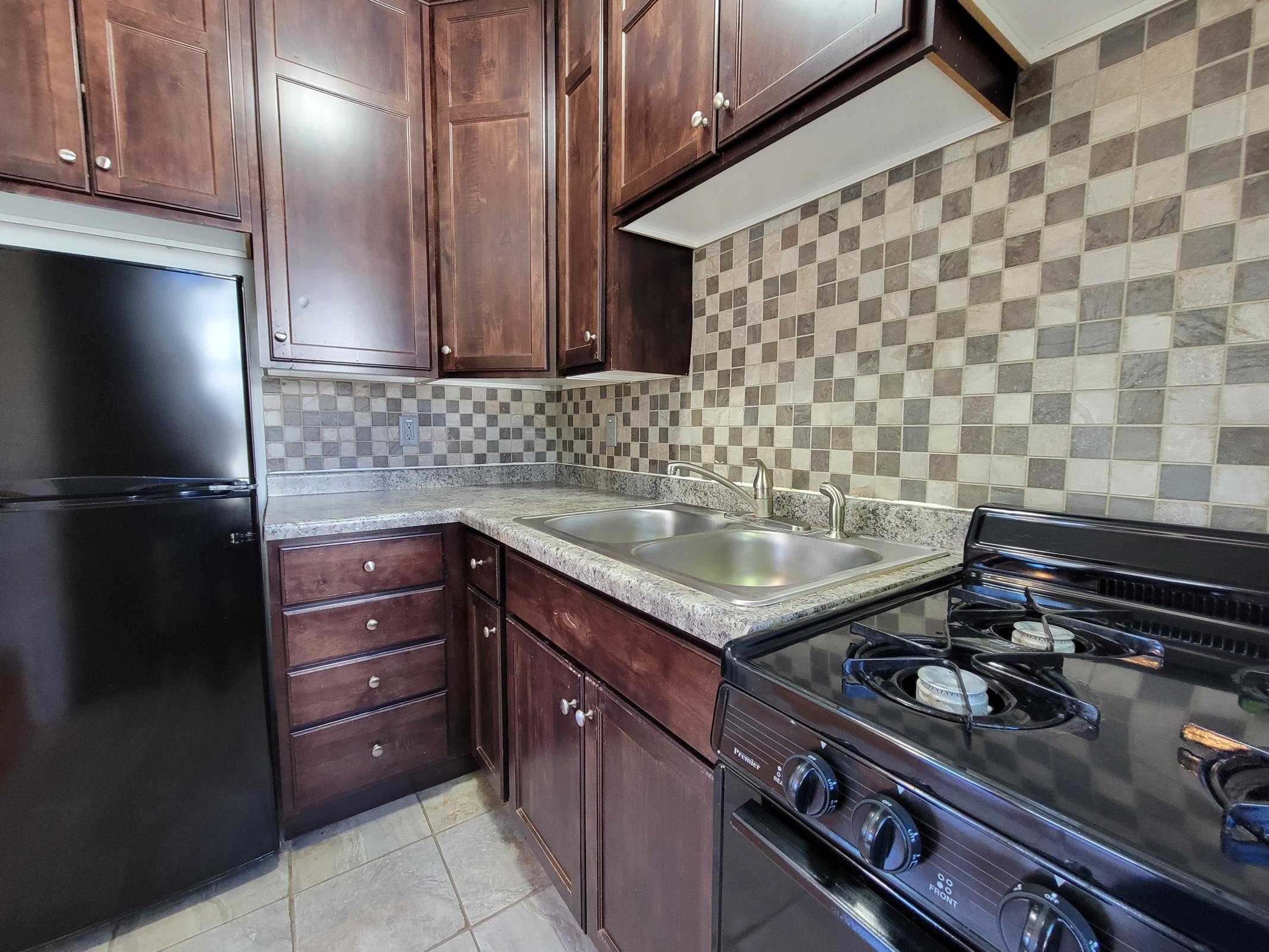 A modern kitchen featuring dark wood cabinets, a black refrigerator, a stainless steel sink with two basins, and a black gas stove. The backsplash consists of a mosaic of gray and beige tiles, complementing the granite countertop and tiled floor.