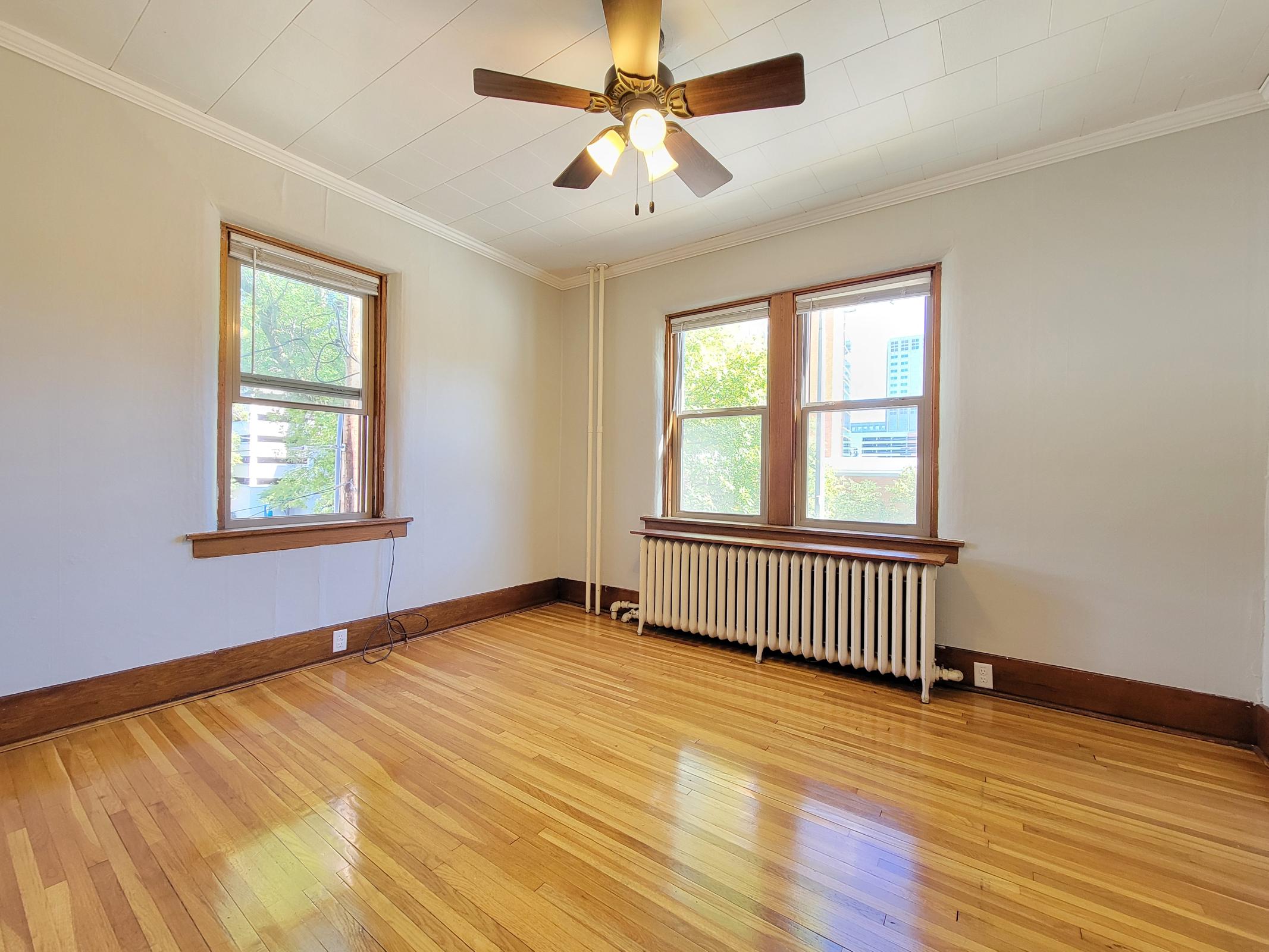 A bright, empty room with hardwood floors, featuring three windows that allow natural light. The walls are painted light gray, and a ceiling fan hangs above. A radiator is along the wall beneath the windows, showcasing a simple, cozy atmosphere.