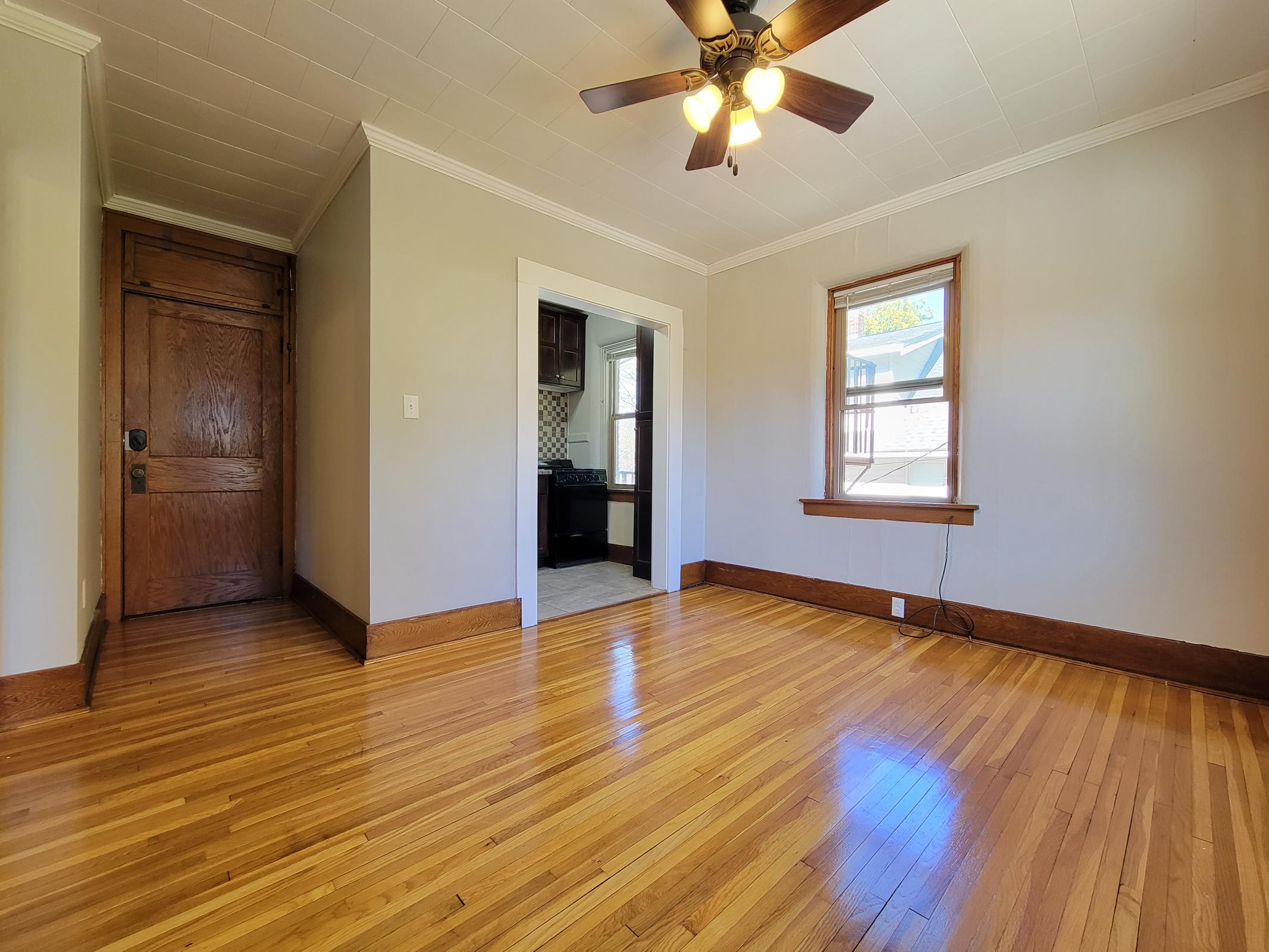 A bright living room featuring hardwood floors, a ceiling fan, and a large window allowing natural light. The walls are painted in a soft gray color, and there's a doorway leading to a kitchen area with dark cabinetry visible in the background. The overall ambiance is warm and inviting.