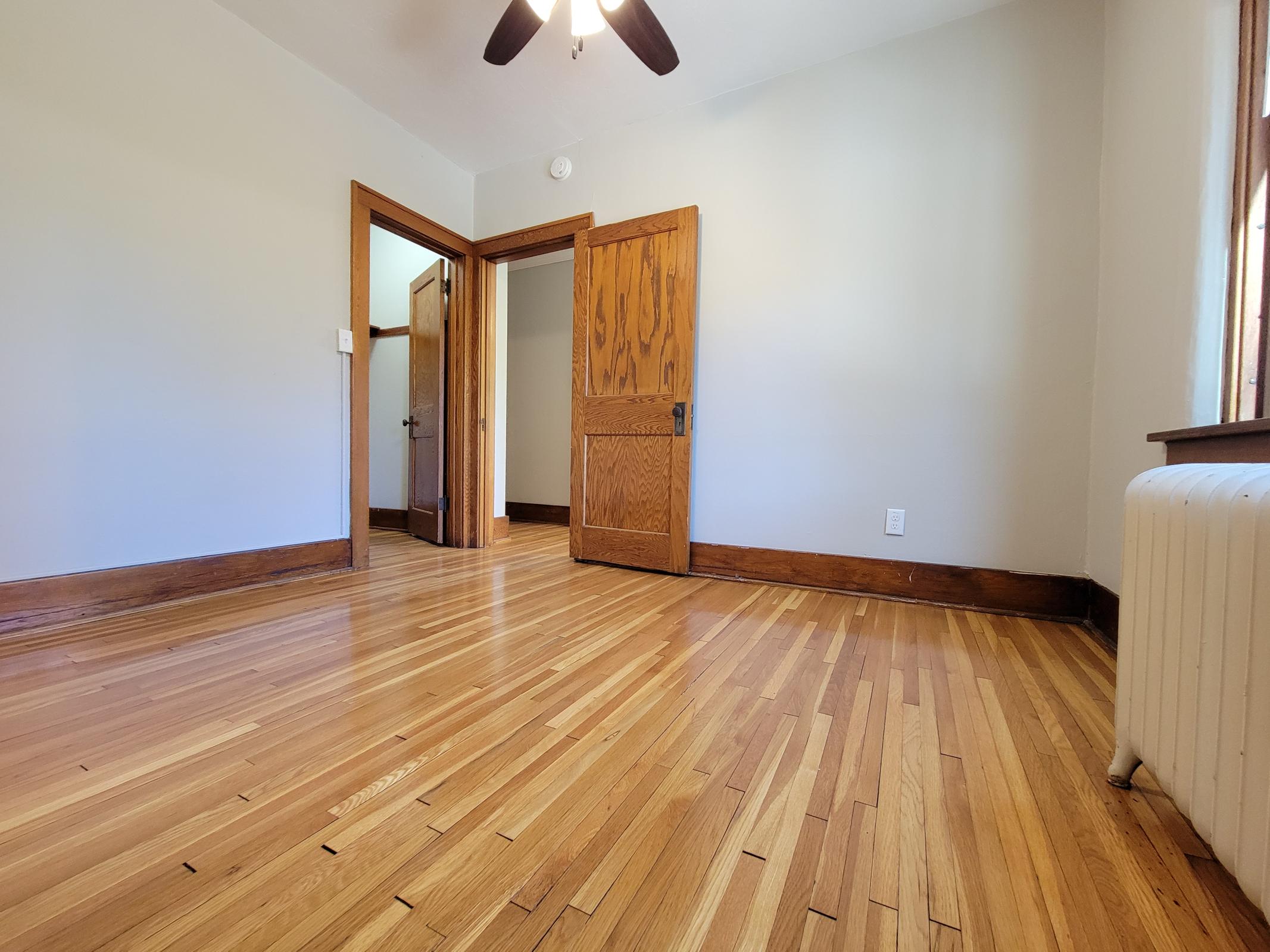 A spacious, empty room featuring hardwood flooring, a ceiling fan, and two wooden doors leading to adjacent rooms. The walls are painted light grey, and a radiator is visible near the window, contributing to a cozy atmosphere.