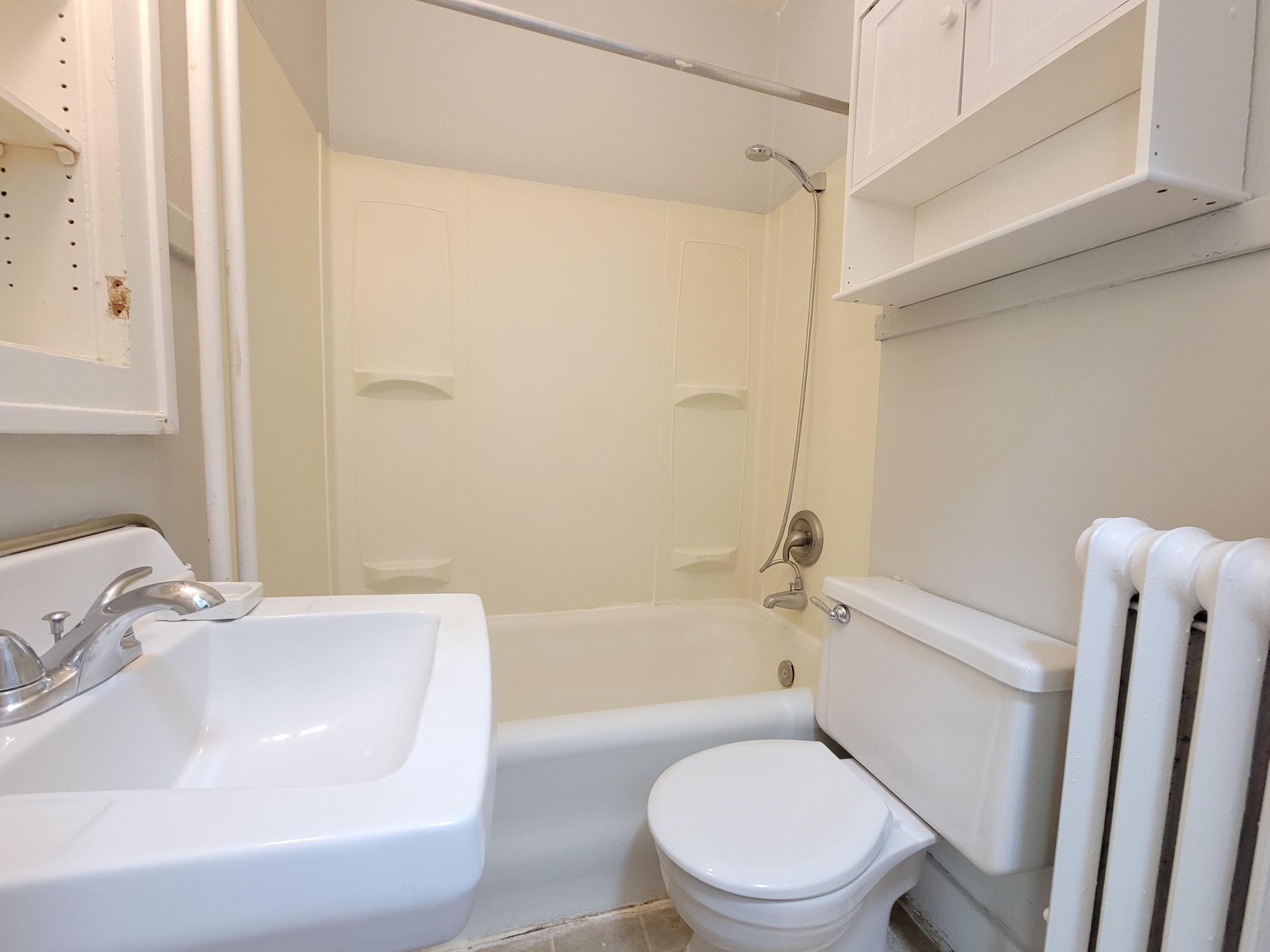 A small bathroom featuring a bathtub with a shower, a sink, a toilet, and a wall-mounted cabinet. The walls are painted a light color, and there is a radiator on the side. The overall design is simple and functional.