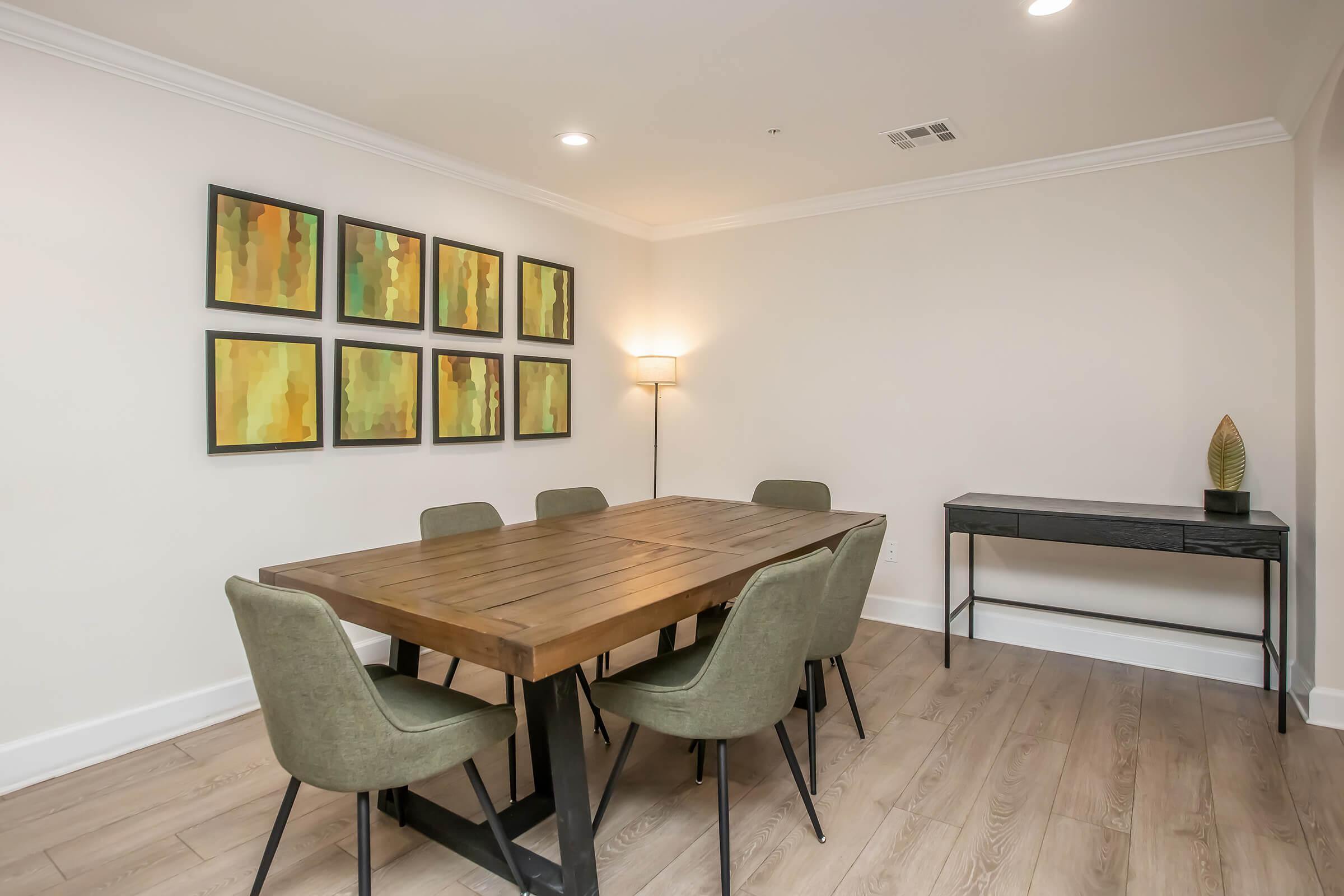 A spacious dining area featuring a wooden dining table surrounded by six green upholstered chairs. On the wall, there are six framed abstract paintings in shades of yellow and green. A modern floor lamp provides ambient lighting, and a slim console table stands against the wall with a small decorative plant.