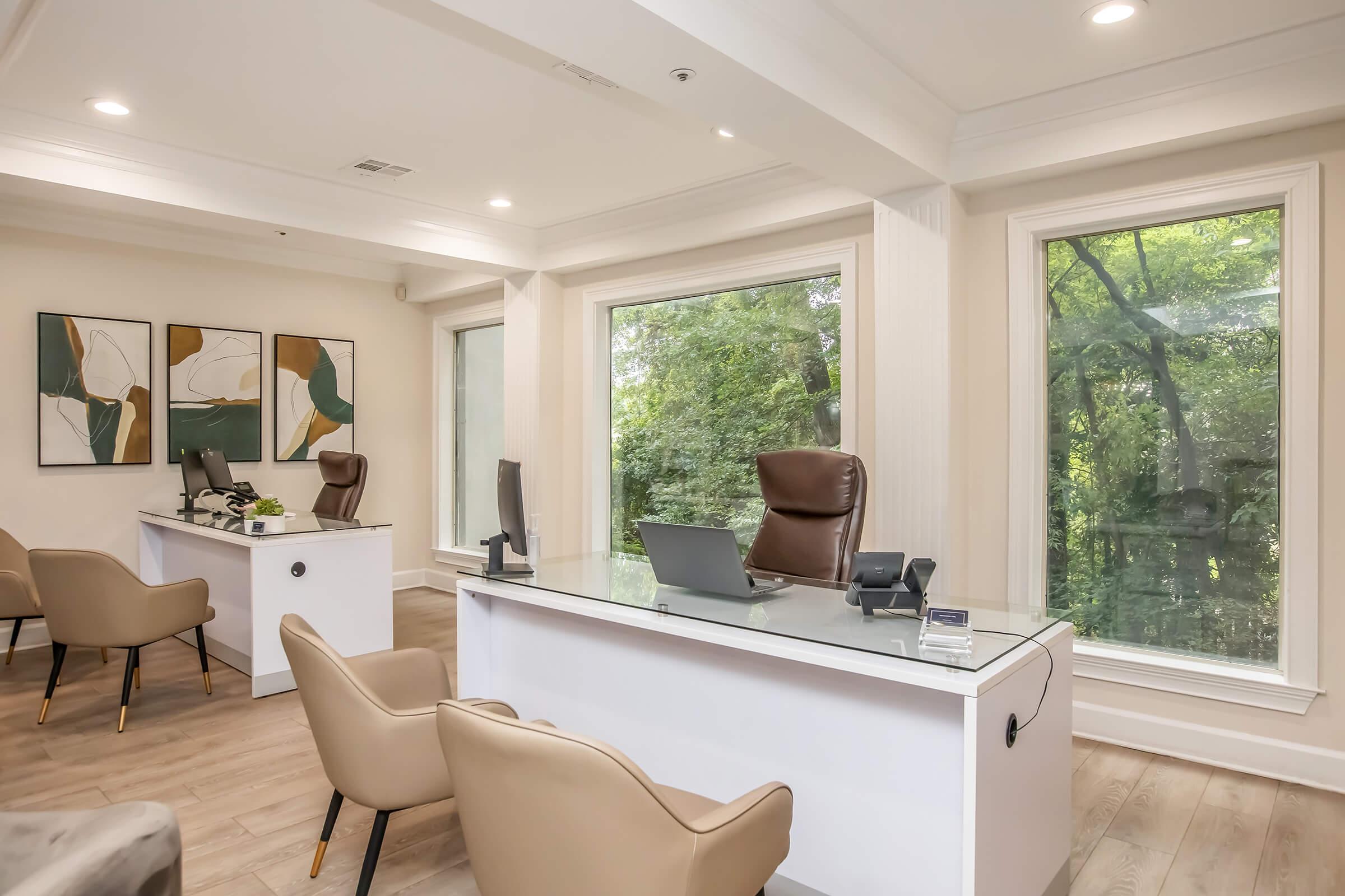 A bright and modern office space featuring a sleek white desk with a computer, a comfortable brown chair, and two additional chairs. Large windows provide a view of lush greenery outside, enhancing the peaceful atmosphere. Abstract art is displayed on the walls, adding a touch of elegance to the space.
