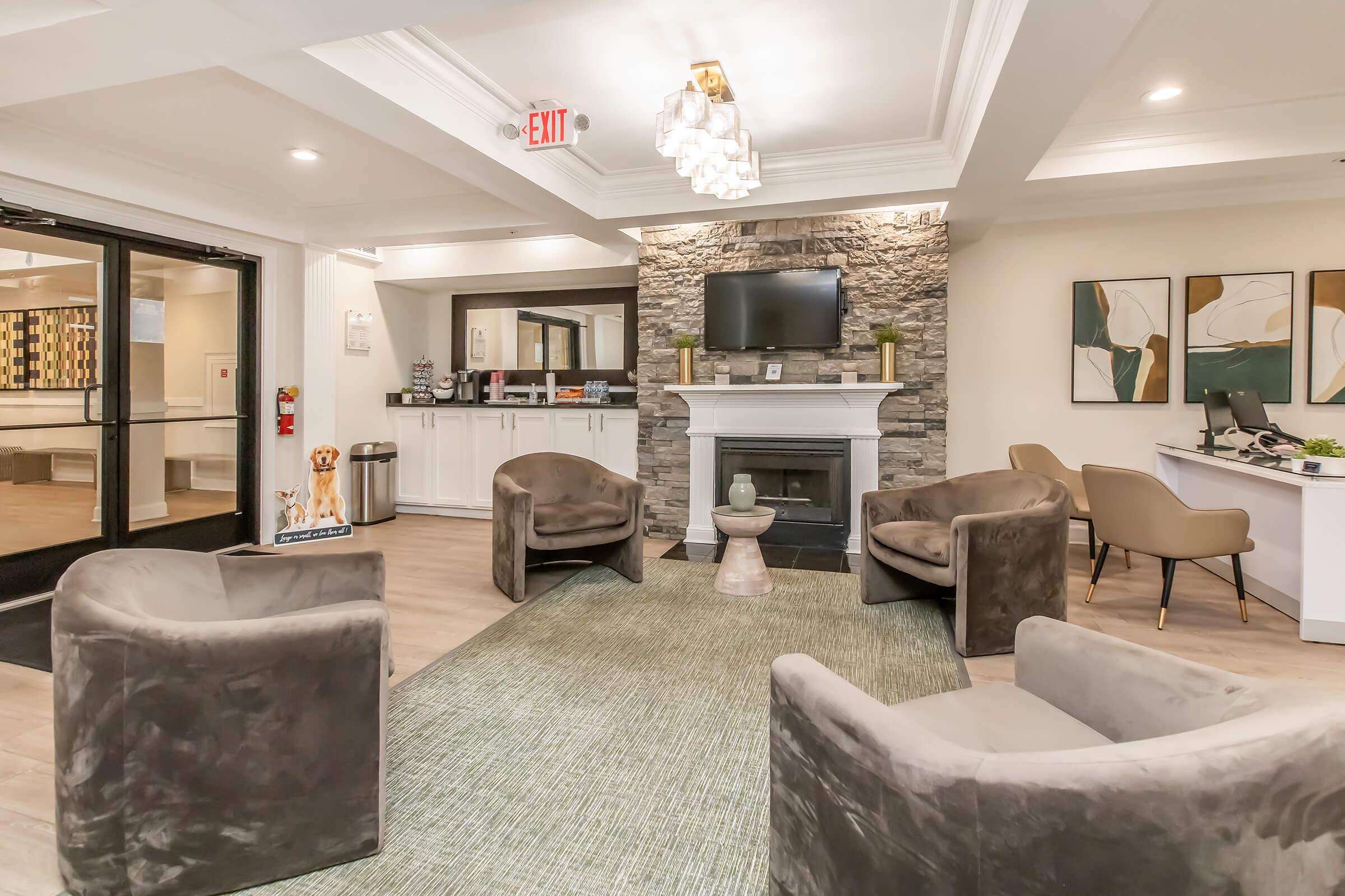 A cozy lobby area featuring a stone fireplace with a television above it. Four plush chairs are arranged around a modern coffee table on a green area rug. To the side, there is a reception desk with a computer and seating, along with a garbage can and decorative artwork on the walls.