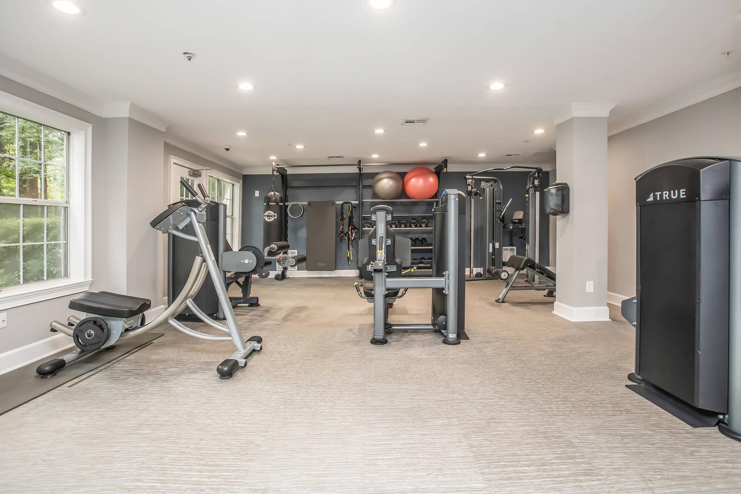 A modern gym interior featuring various exercise equipment including a treadmill, multi-gym, stationary bike, and free weights. The spacious area has large windows allowing natural light, with a neutral color scheme and rubber flooring, creating an inviting workout environment.